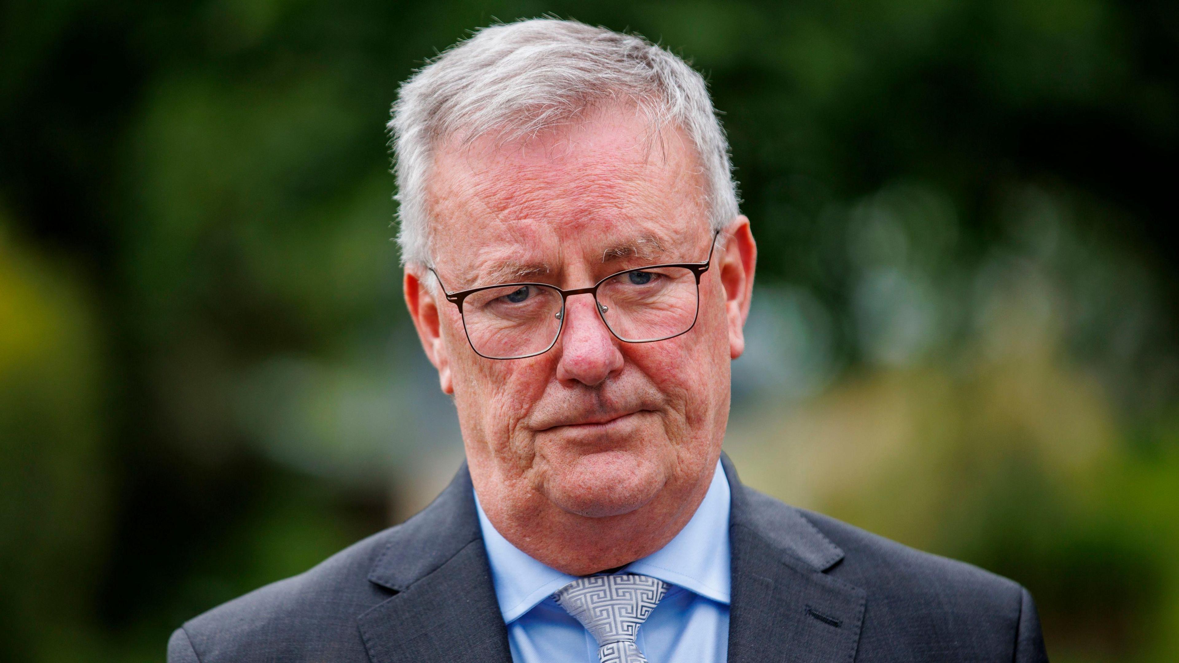 Mike Nesbitt, he is wearing a dark grey blazer with a blue shirt and a white and grey pattern tie. He has white hair and is wearing glasses.