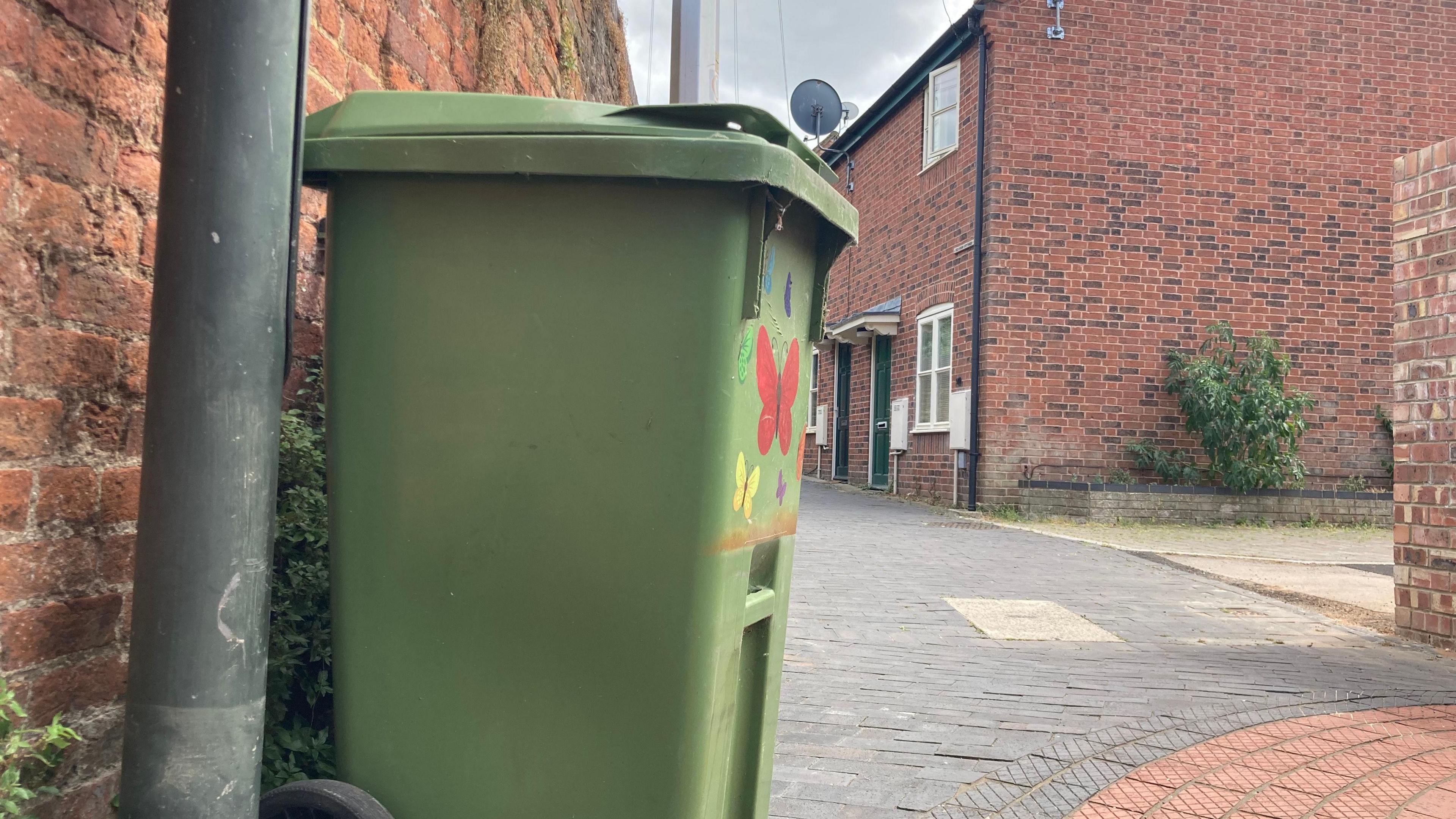 A photo showing a green wheelie bin on a street next to a red bricked wall and a lamp post. In the background there is a row of red brick houses.