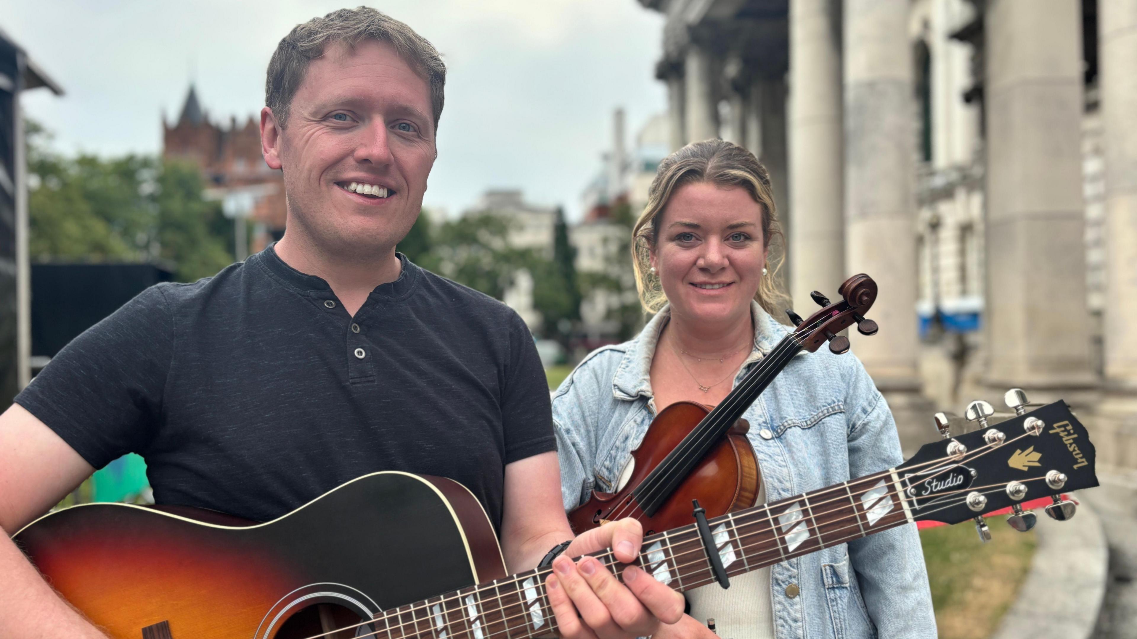 A man and a woman smiling at the camera. The man is holding a guitar and the woman is holding a fiddle. They are standing outside near some large stone pillars. The man is wearing a black t-shirt and the woman is wearing a denim jacket. 