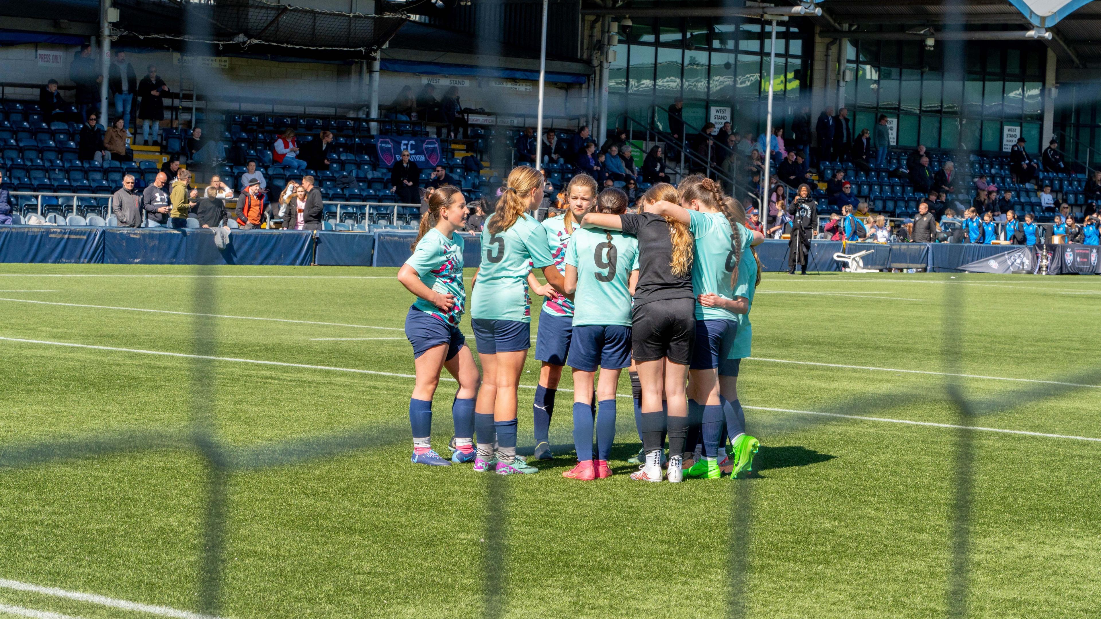 A small group of girls in aquamarine coloured jerseys huddling on a football pitch