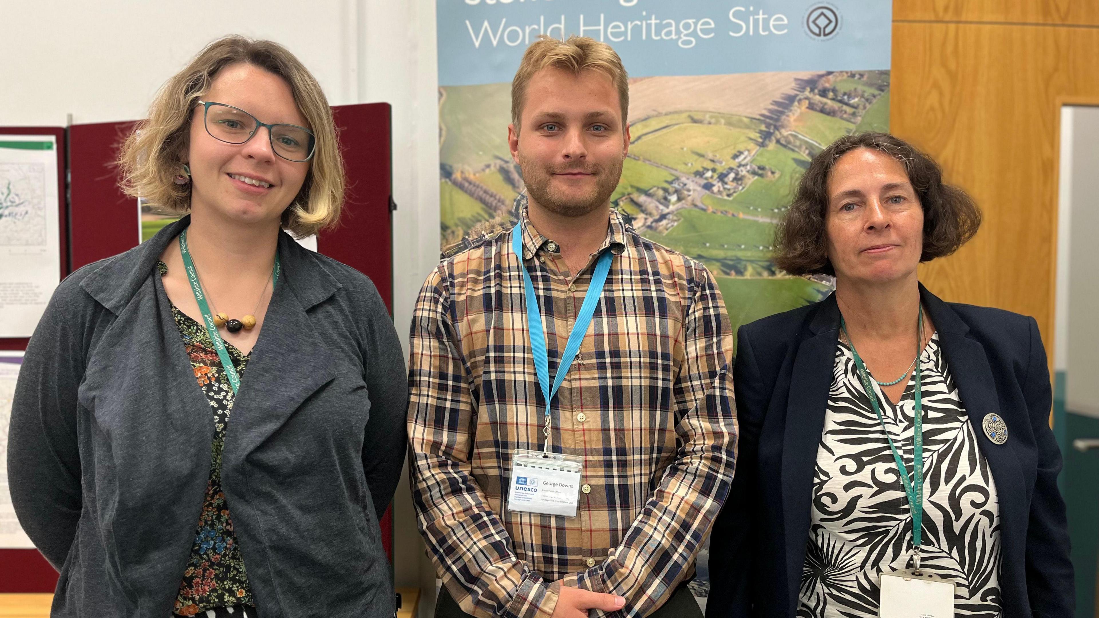 Claire Selman, George Downs and Melanie Pomeroy-Kellinger stand together smiling at the camera in front of banners with pictures of Neolithic monuments