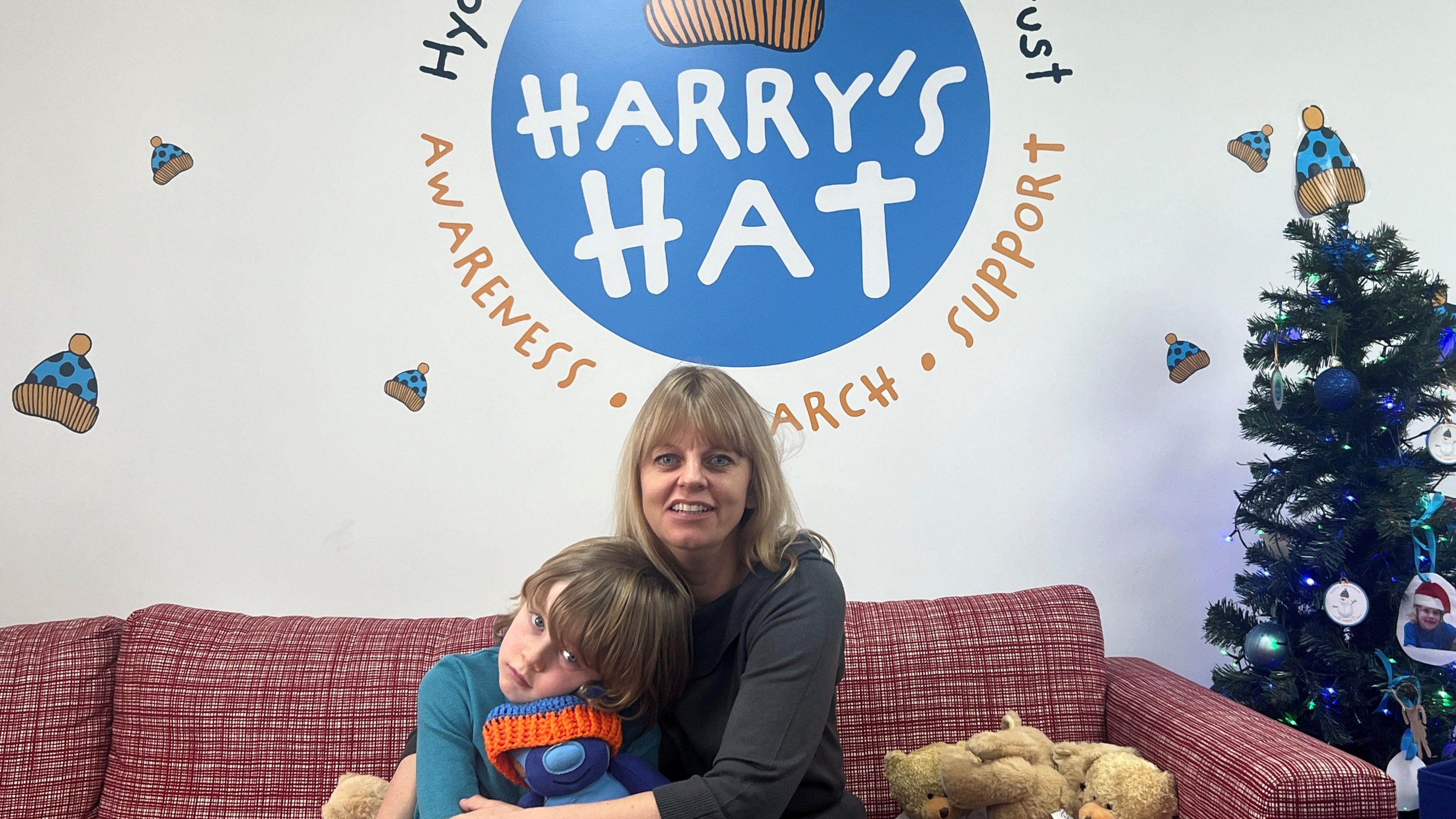 Woman with blonde hair hugging her son in a blue top, who's hugging a blue teddy bear in front of a logo that shows the charity's name, Harry's Hat.