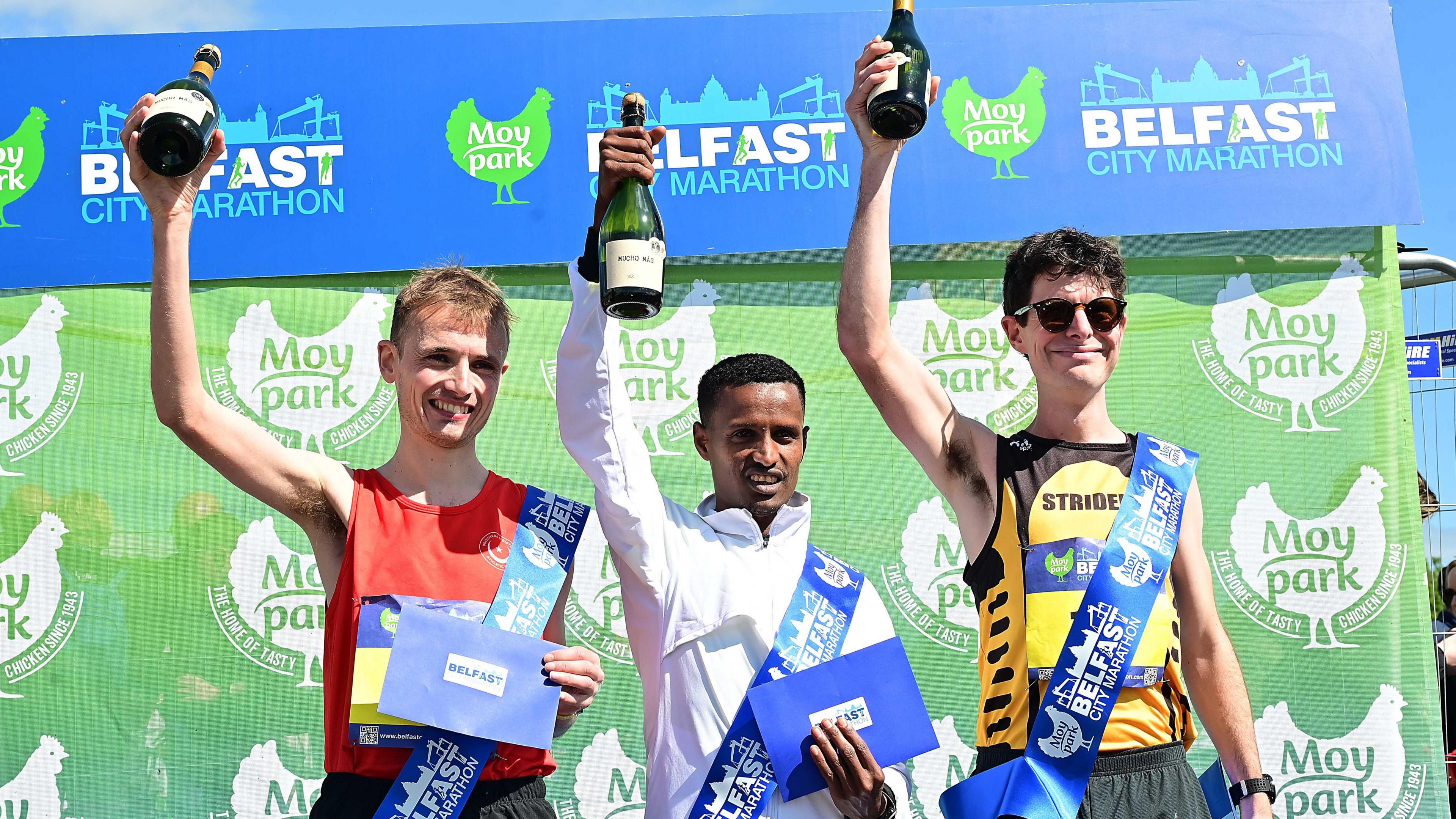 The three male winners of the Belfast Marathon hold up bottles of champagne in one hand and a blue envelope in the other. On the left is runner-up Aaron Hanlon, who is wearing a blue sash and a red athletic top and is smiling. In the middle is Tilahun Nigussie, who is wearing a white zip up jacket and is smiling at the camera. On the right is Stephen Connolly, wearing a yellow and black athlete best with 'Strident' written across the chest.