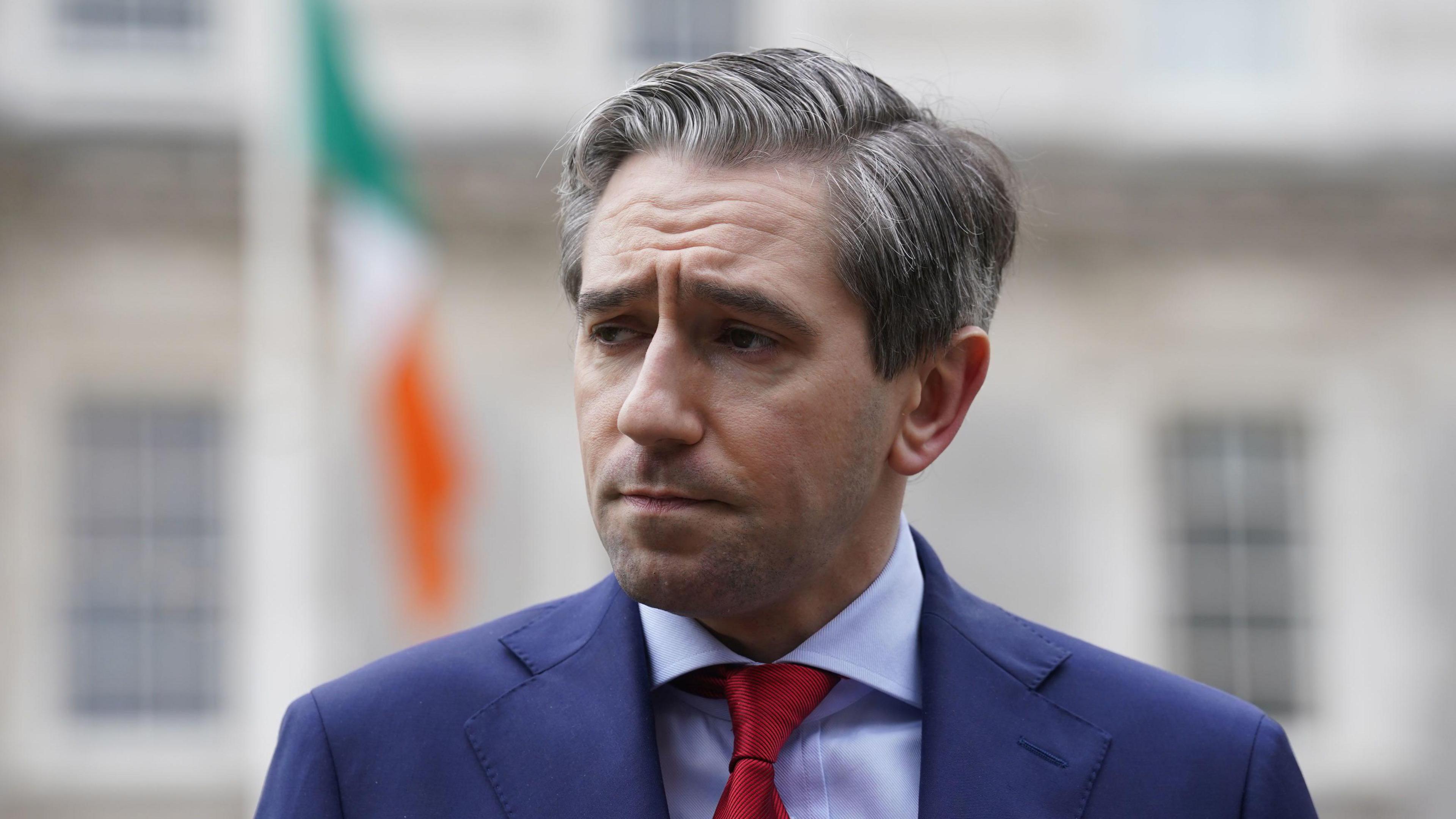 Tánaiste Simon Harris, pictured from the chest up, wearing a navy suit, light blue shirt and red tie. He has greying short hair. There is a blurred grey building and Irish flag in the background. 