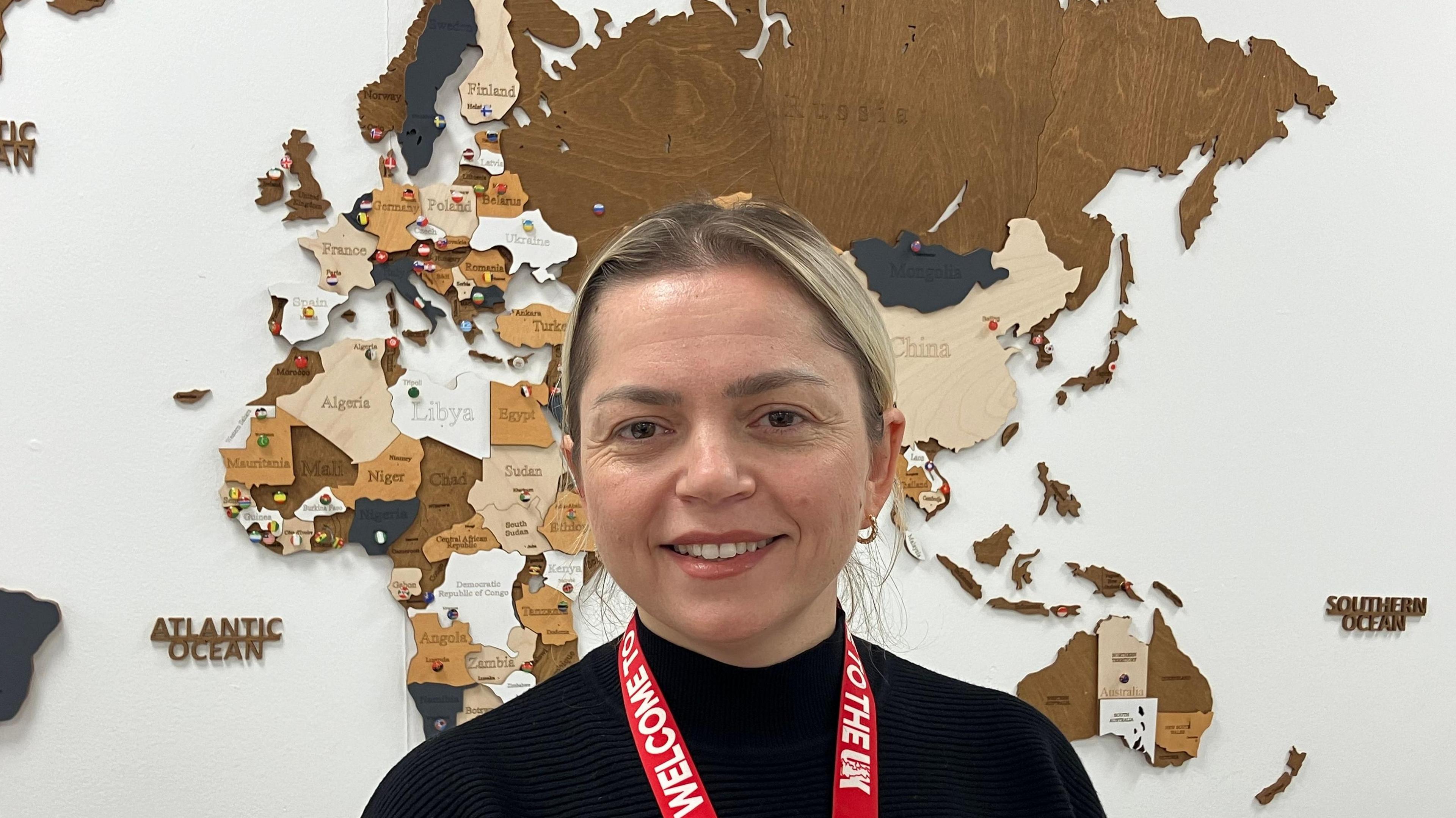 A head and shoulders picture of Ilda Sinani in front of a world map. She is wearing a black jumper and has blonde hair tied back from her face.