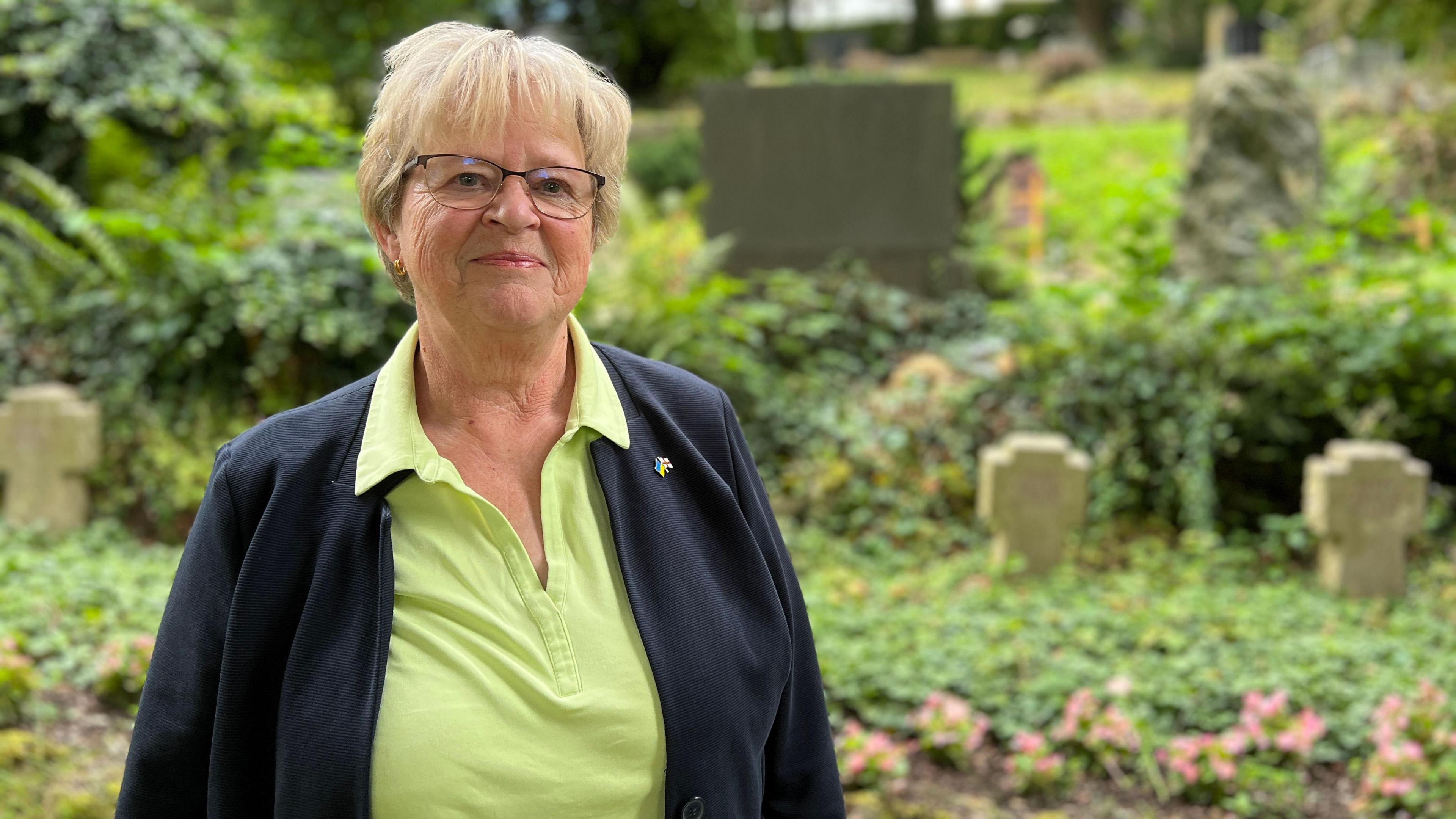 Helga has short, light-colored hair is standing outdoors in a garden or park area. She is wearing a light green collared shirt and a dark jacket. In the background, there are various plants, trees, and what appear to be gravestones or memorial markers.
