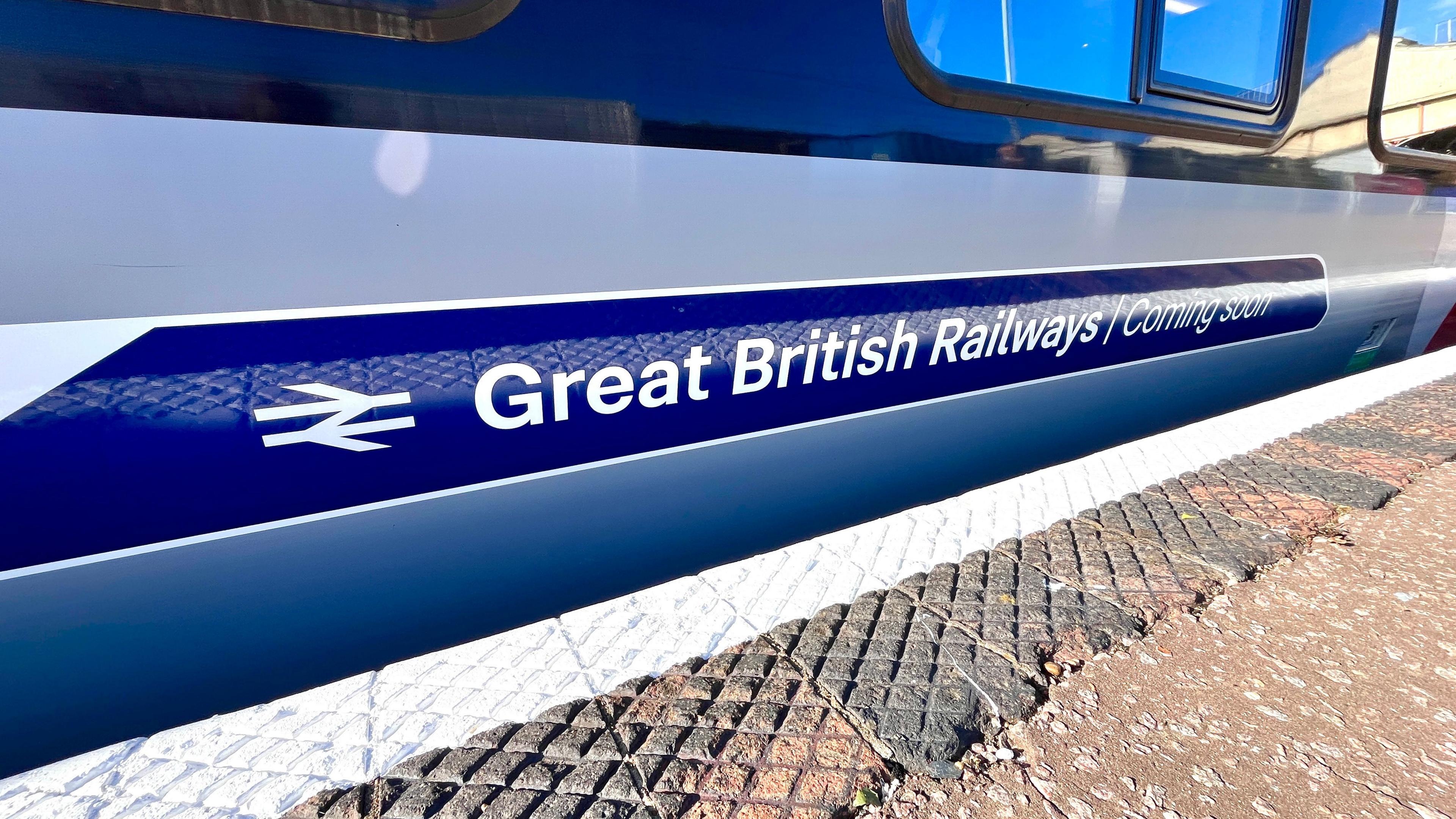 An image of the side of a train, standing at a platform. Only the lower part of a carriage can be seen, along side the edge of the platform. A sticker on the side has the parallel lines railway logo and the text 'Great British Railways - Coming Soon'. The lower section of two windows are also visible above and to the right of the sticker. A thick white line denotes the edge of the platform, which is otherwise covered in patterned tiles and light coloured asphalt.