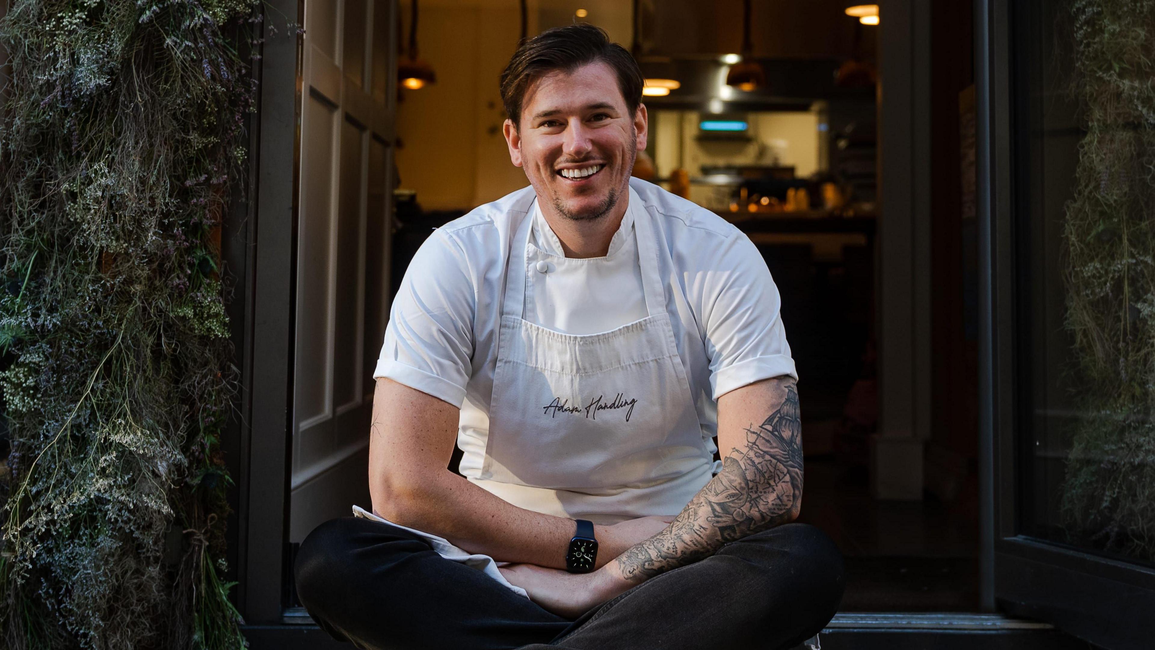 Adam Handling in a white t-shirt, black trousers and white apron. He sits cross legged on the floor outside a restaurant's front door.