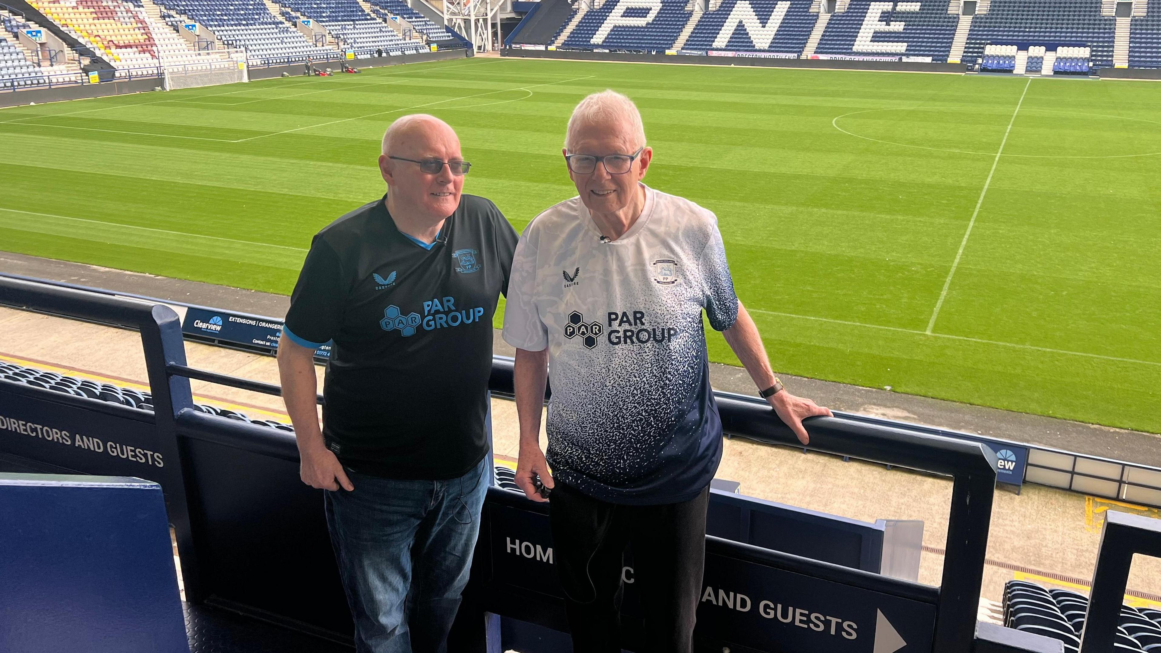 Peter and Terry Kilgariff are standing side by side in the stand at Deepdale Stadium - the home of Preston North End. They are both wearing Preston North End tops and the pitch can be seen behind them.