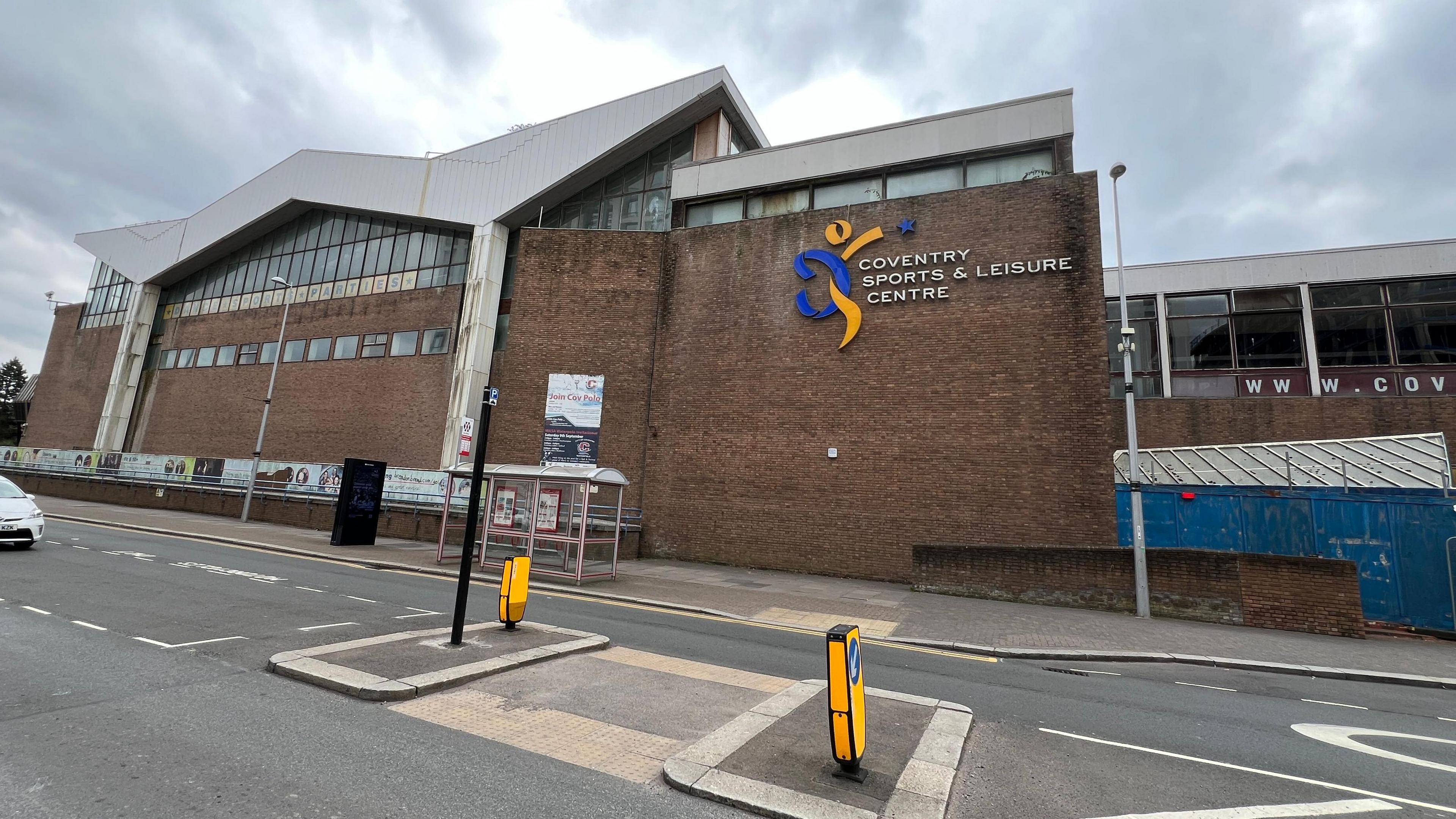 Outside view of Coventry Sports and Leisure Centre on Fairfax Street. A road runs in front of a brick building with large glass windows at the top and a jagged roof. The sign identifying the building as 'Coventry Sports and Leisure Centre' is on the brick wall.