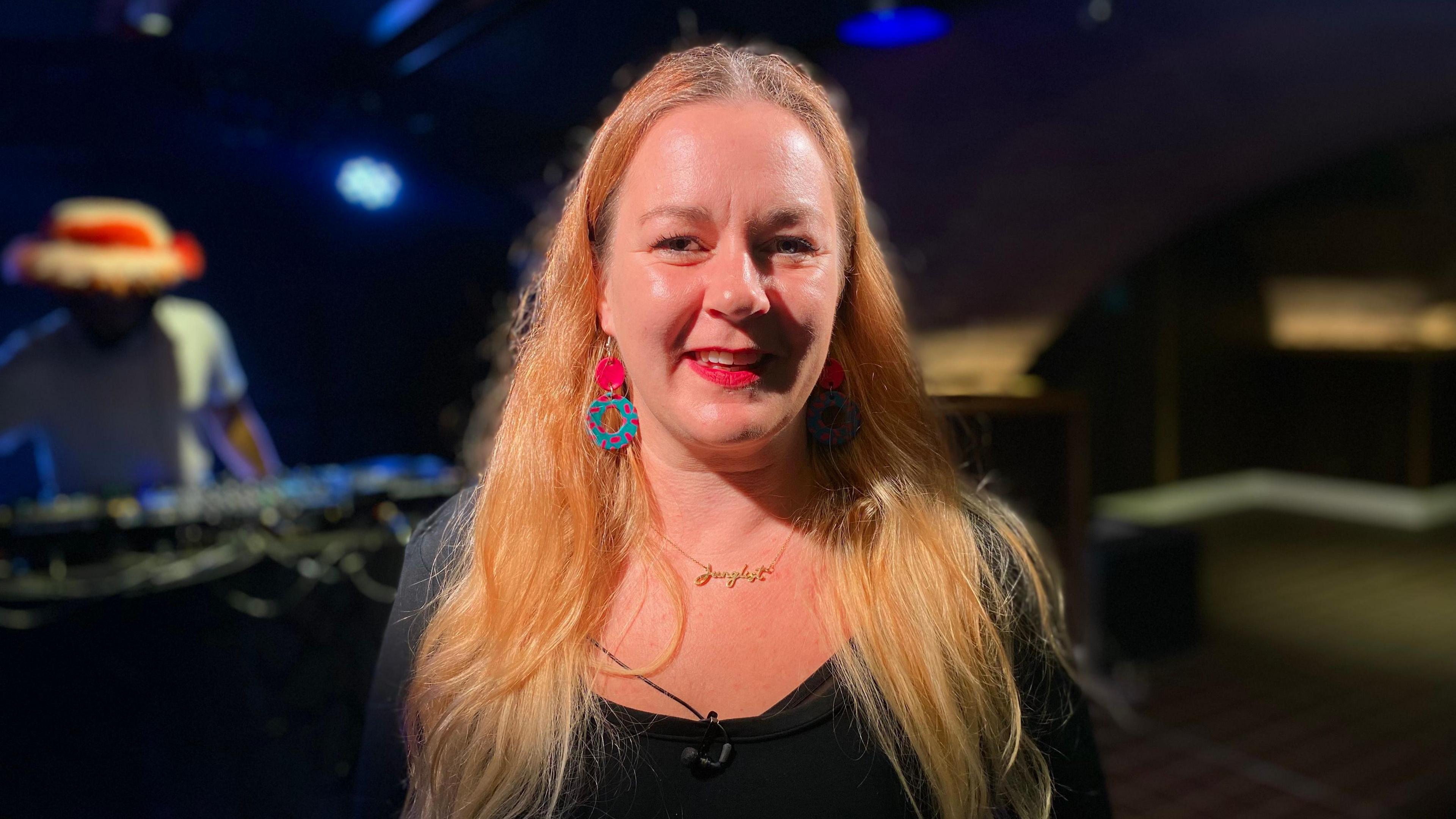 Head and shoulders image of Carly Heath, who has long blonde hair, wearing a black top and brightly coloured earrings, standing in a dark room