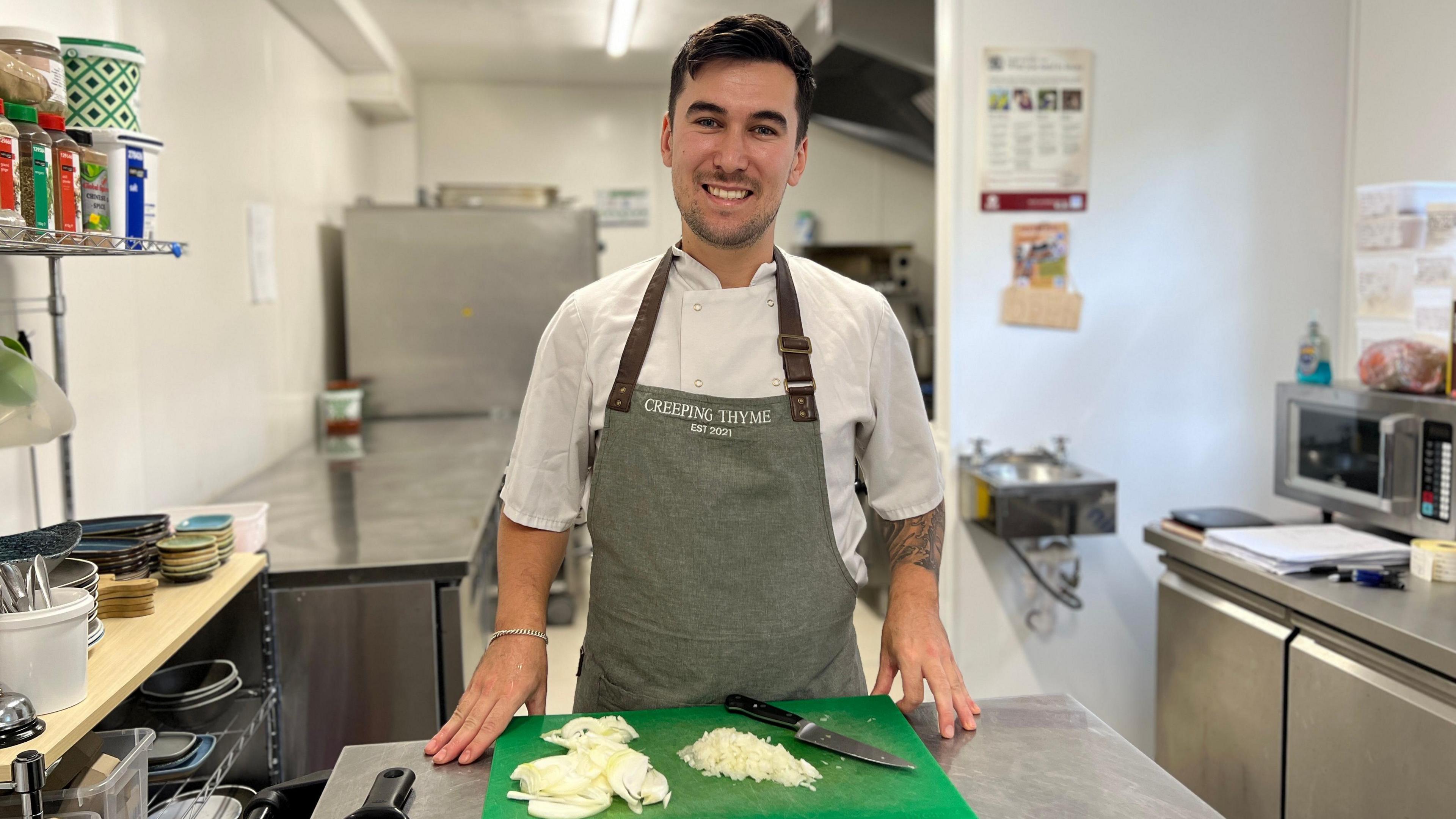 Lewis is wearing chef whites with a grey apron with brown leather straps on top bearing the words 'Creeping Thyme, est 2021' on the front. He is smiling at the camera standing in front of a green chopping board with chopped white onion on it and a knife. In the background and to the sides parts of a professional stainless steel kitchen are visible.