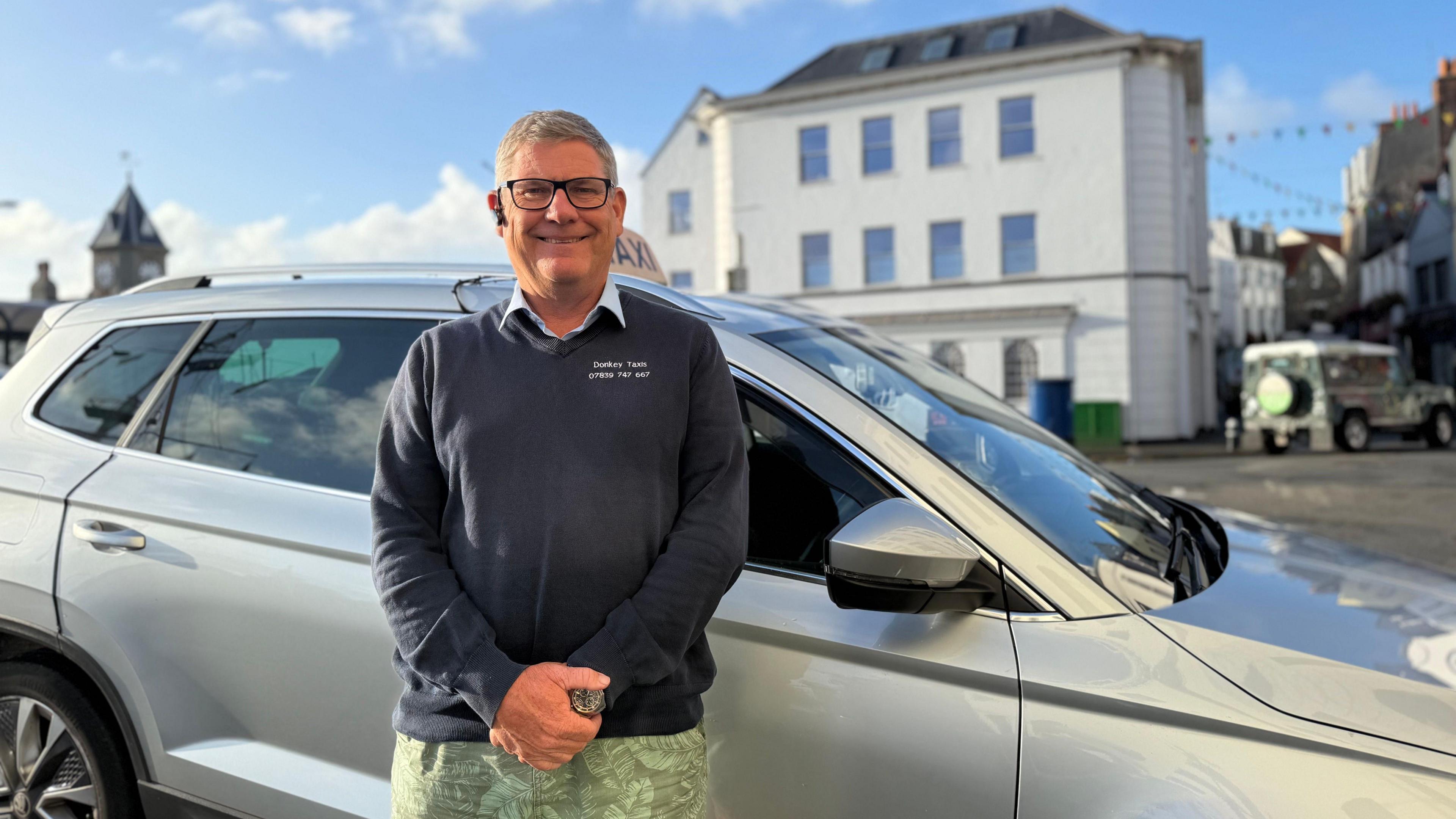 Photo of a man standing in front of a silver taxi. He is wearing a navy jumper with a blue collar poking out the top. He has short grey hair and has black glasses on