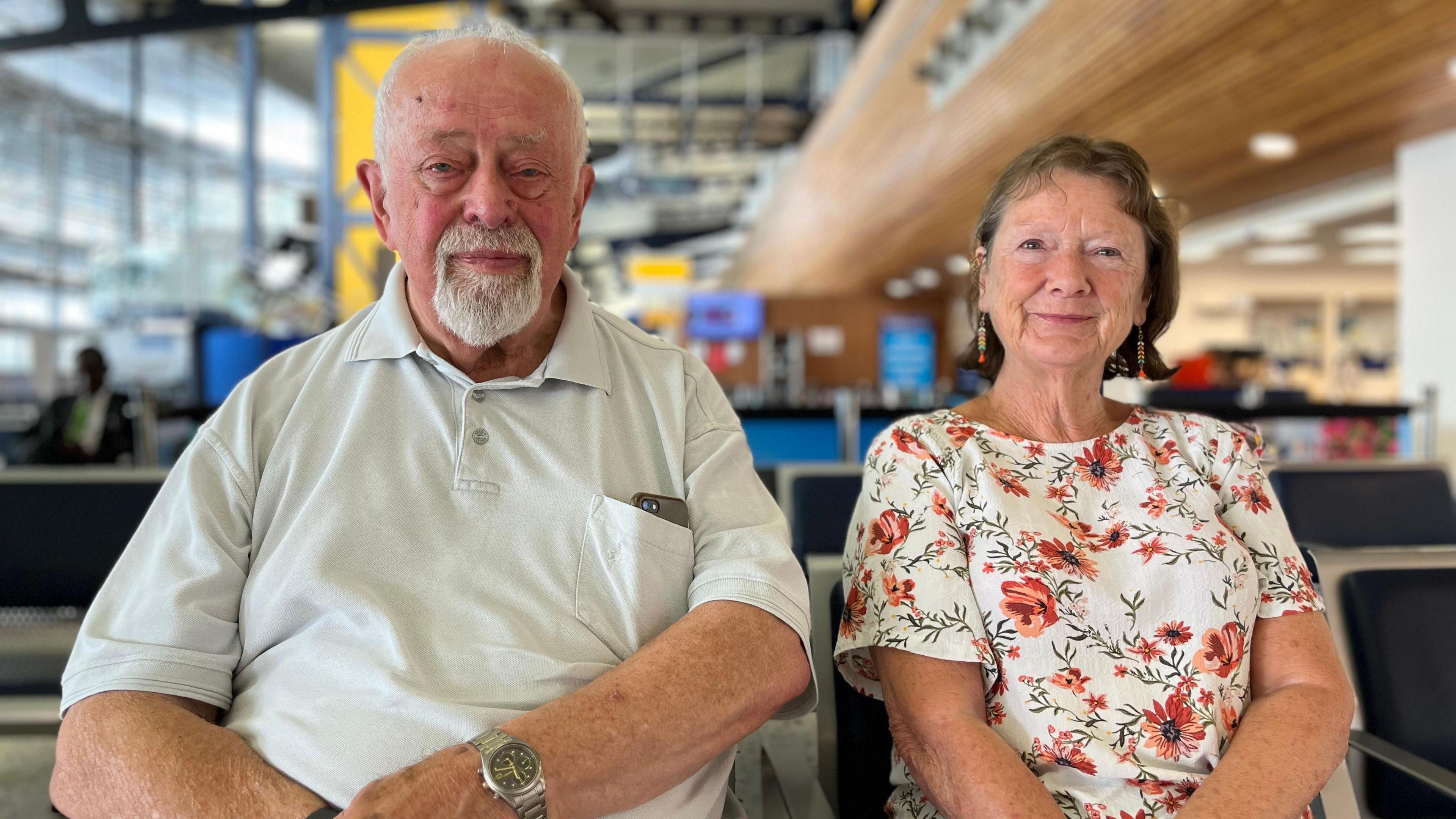 Peter and his wife Val are sitting down on the airport waiting seats. Peter has short white hair and goatie beard. He is wearing a creme t-shirt. Val has shoulder length dark brown hair. She is wearing a white top with red flowers scattered on it.