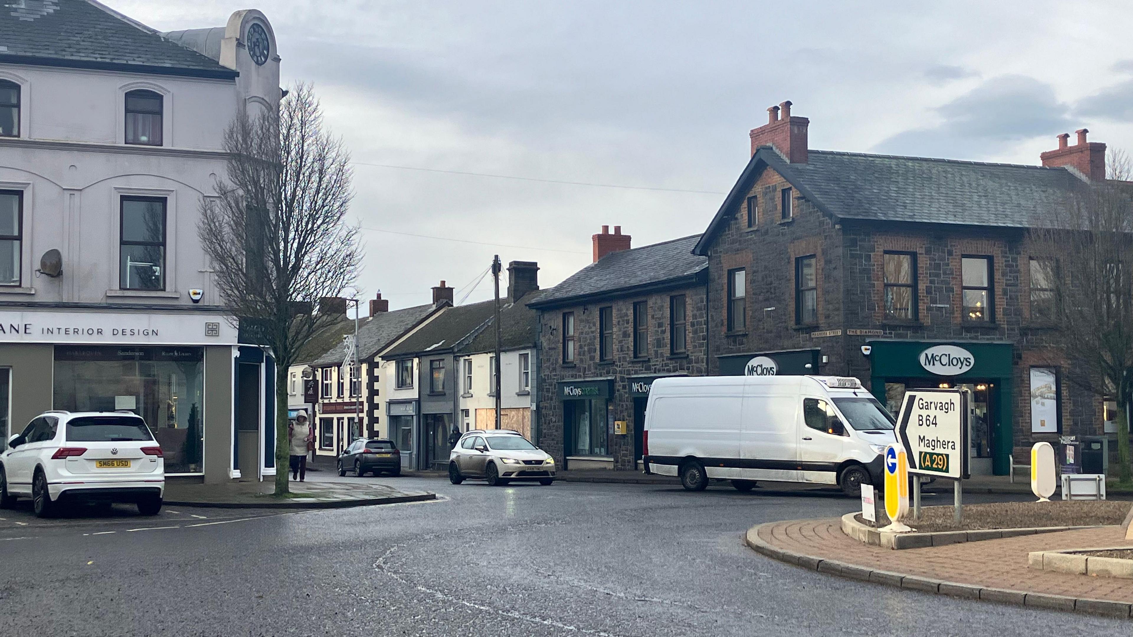 Roundabout in Kilrea centre. Three cars and a van can be seen. A sign on the roundabout shows directions for Garvagh and Maghera.