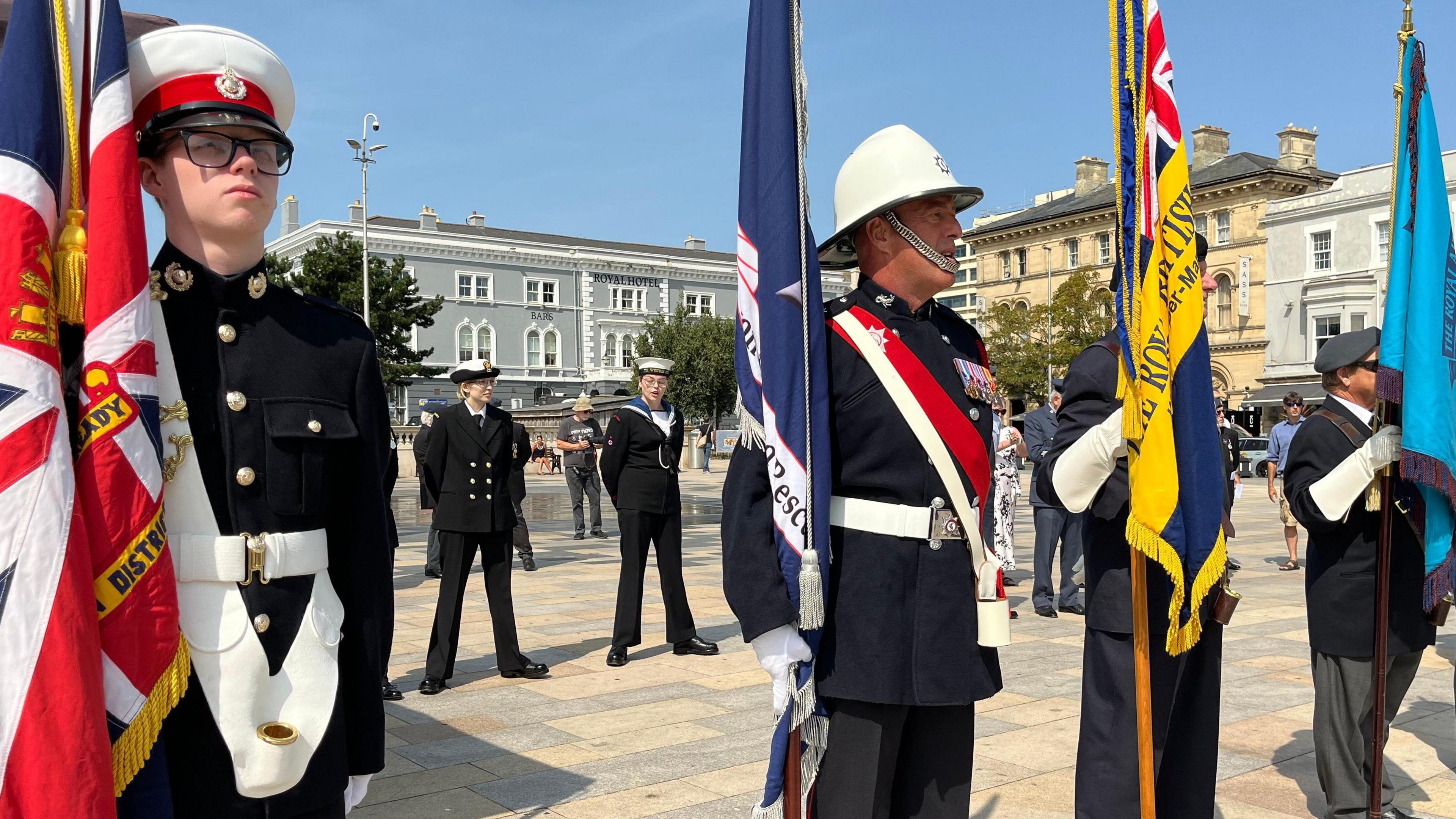 Four men in military uniform stand in a line in a town square, each holding a different flag. Members of the Navy can be seen standing behind them. There are grand buildings at the edge of the square.