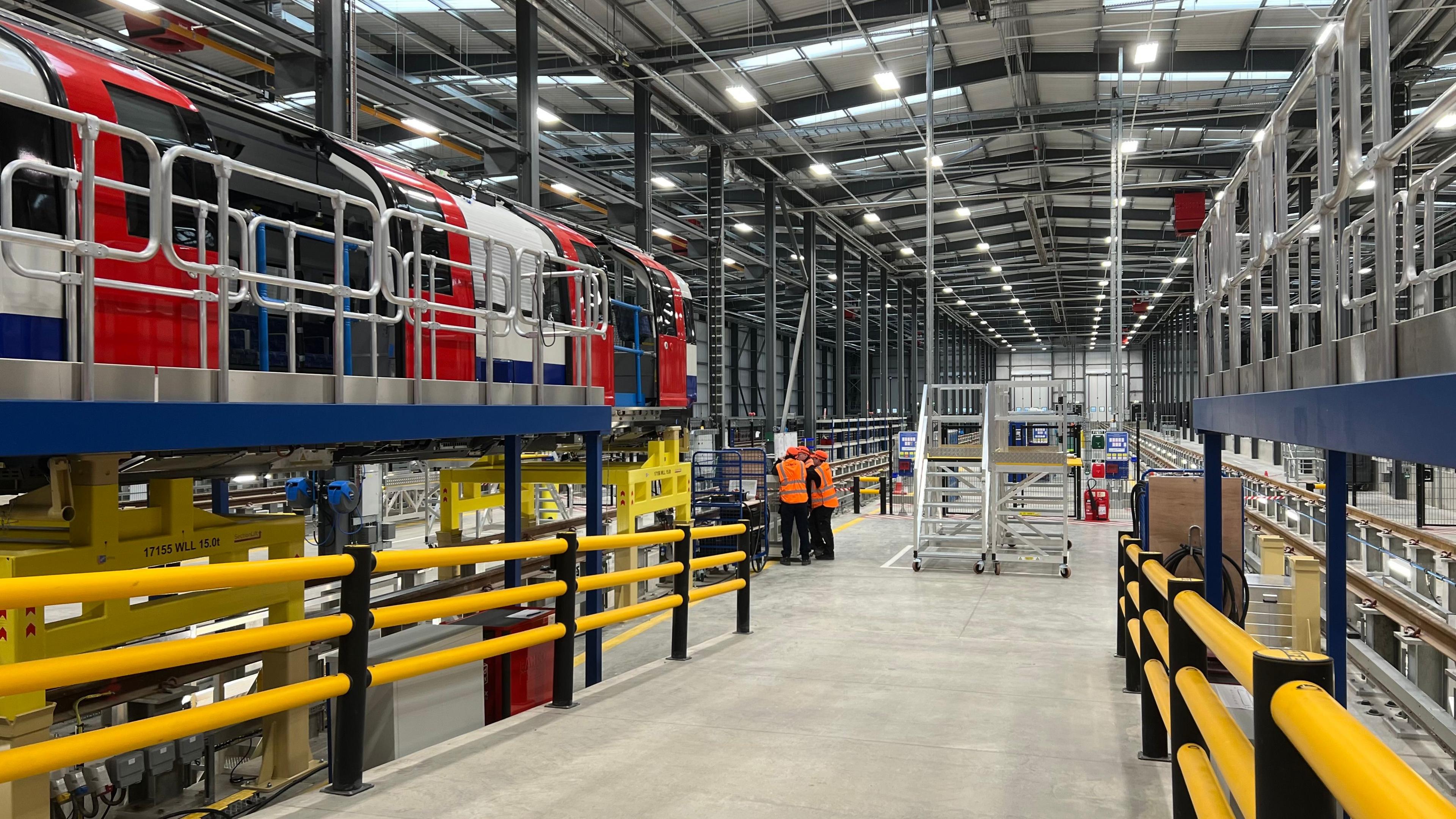 A view inside the factory. A ramp leading down to the main floor has yellow rails either side. To the left is a red, white and blue tube carriage propped up on a stand and three workers wearing orange hi-vis vests are talking in the background.