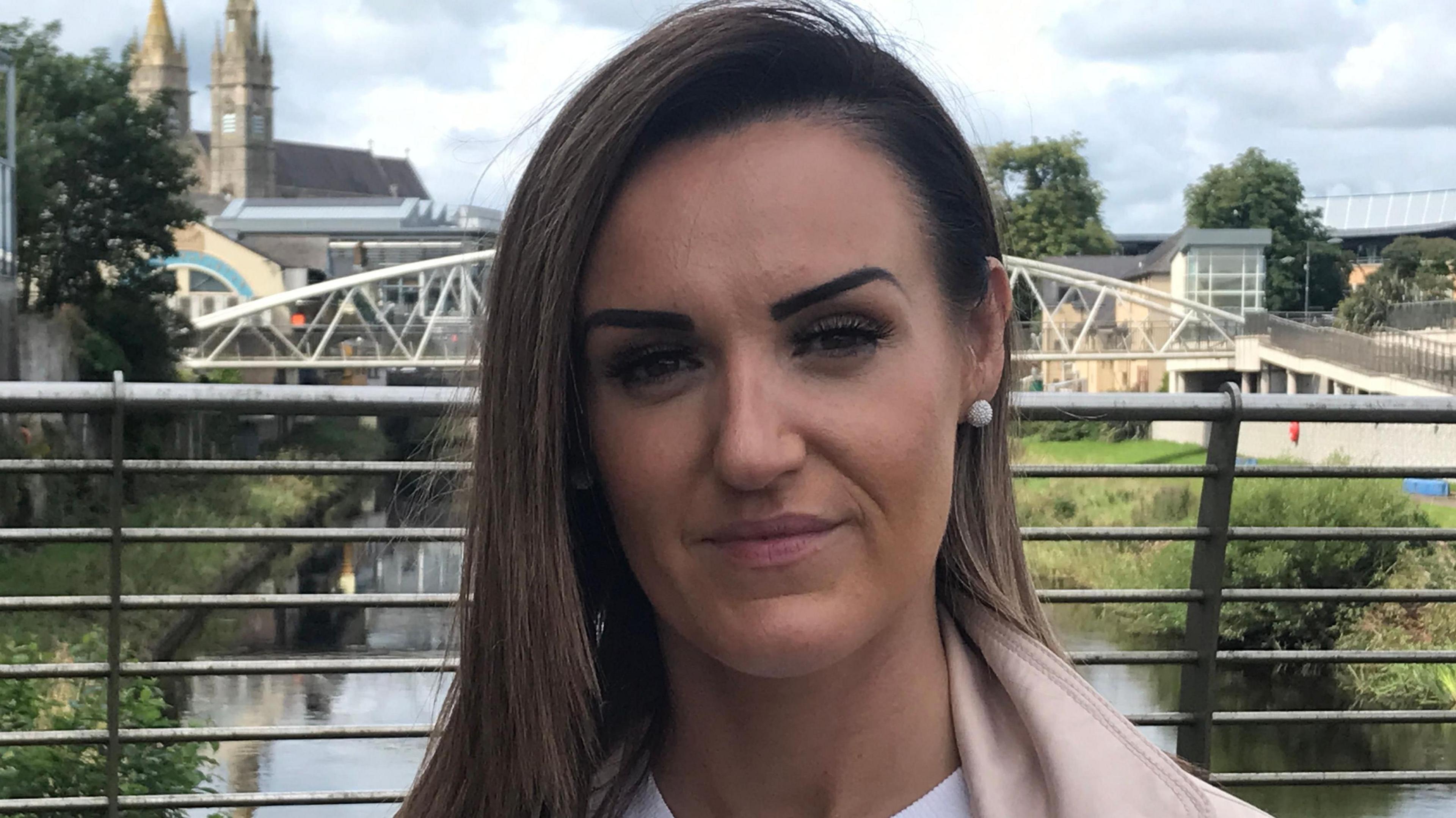 Claire has brown hair and brown eyes. She is wearing a cream coat with a white top. She is standing on a silver bridge, behind her there are buildings and a river.