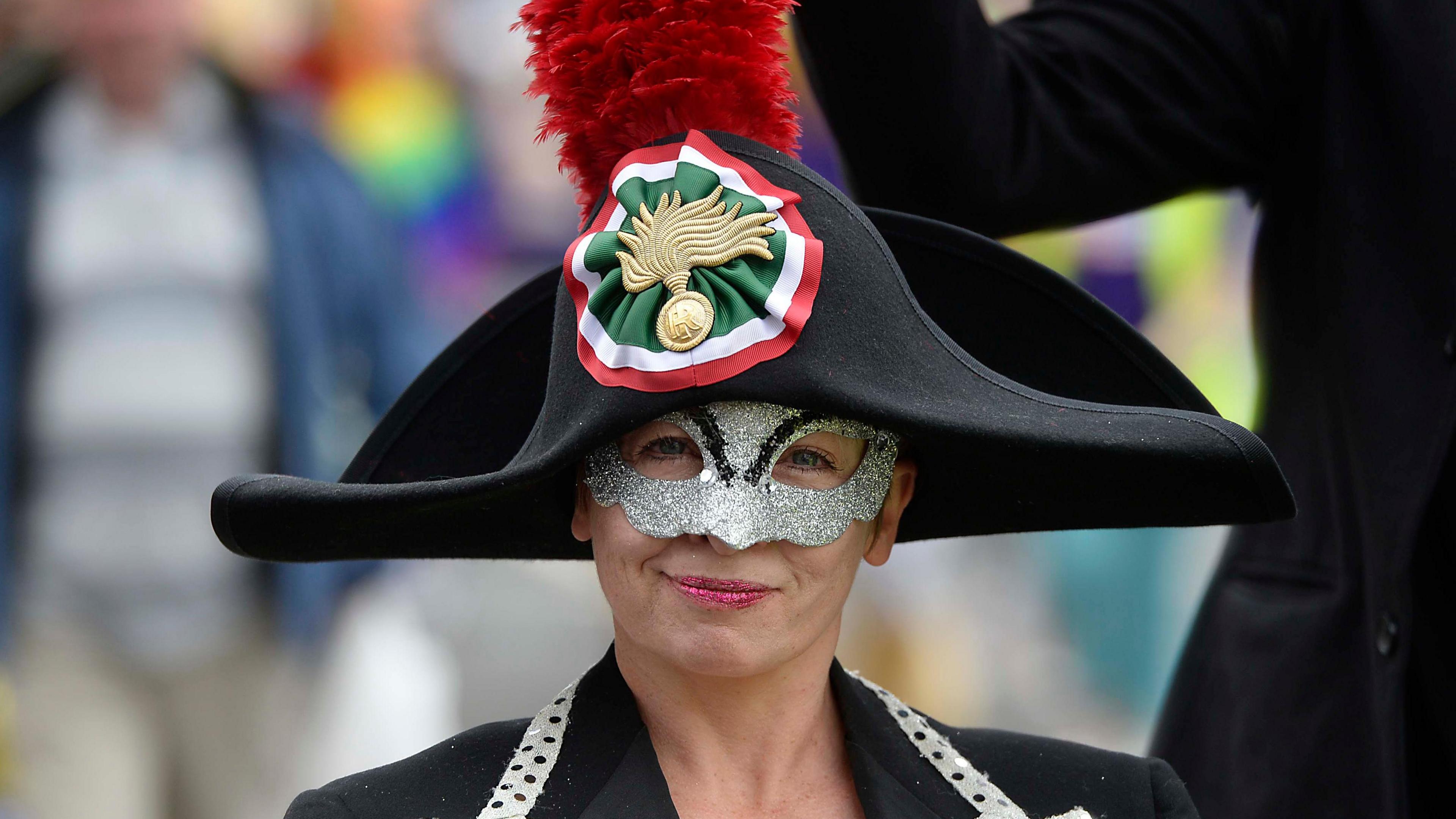 A reveller wearing a silver mask and dressed in a black pirate hat with insignia in green, white and red. A red feather is attached to the hat. 