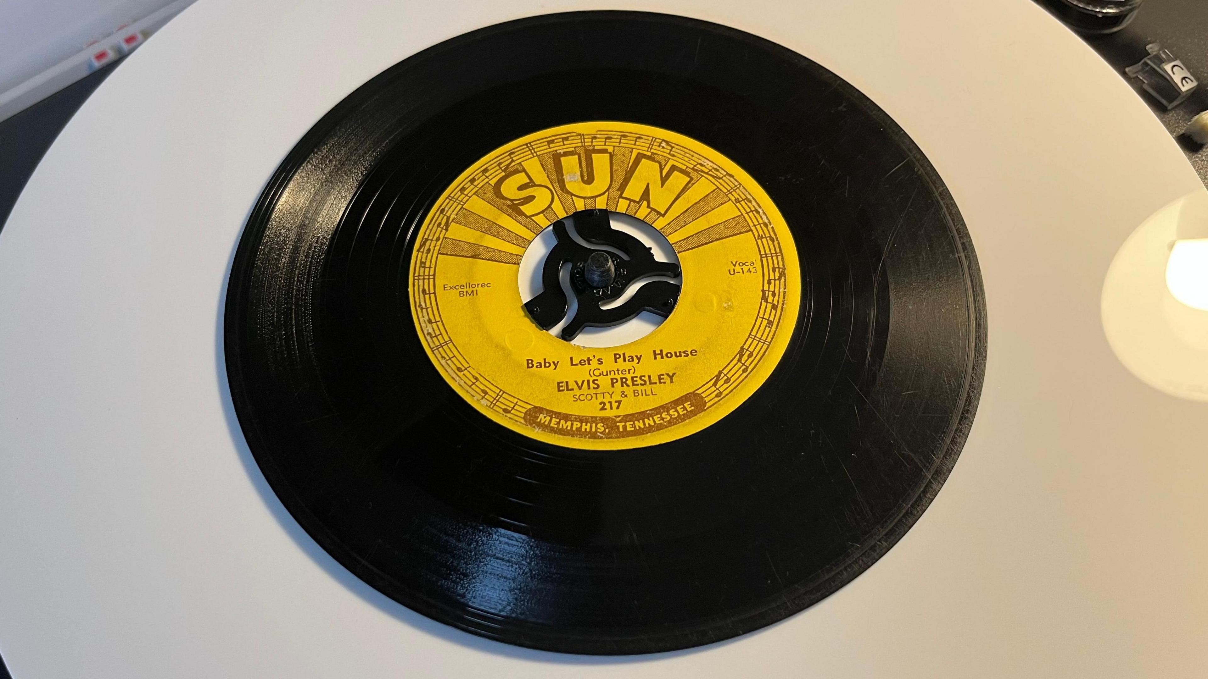 An original Sun Records 45 rpm vinyl record on a turntable. The label on the disc is yellow background with light brown writing it is a song called "Baby Let's Play House" from 1955 and was recorded by Elvis Presley at the Sun studio in Memphis.