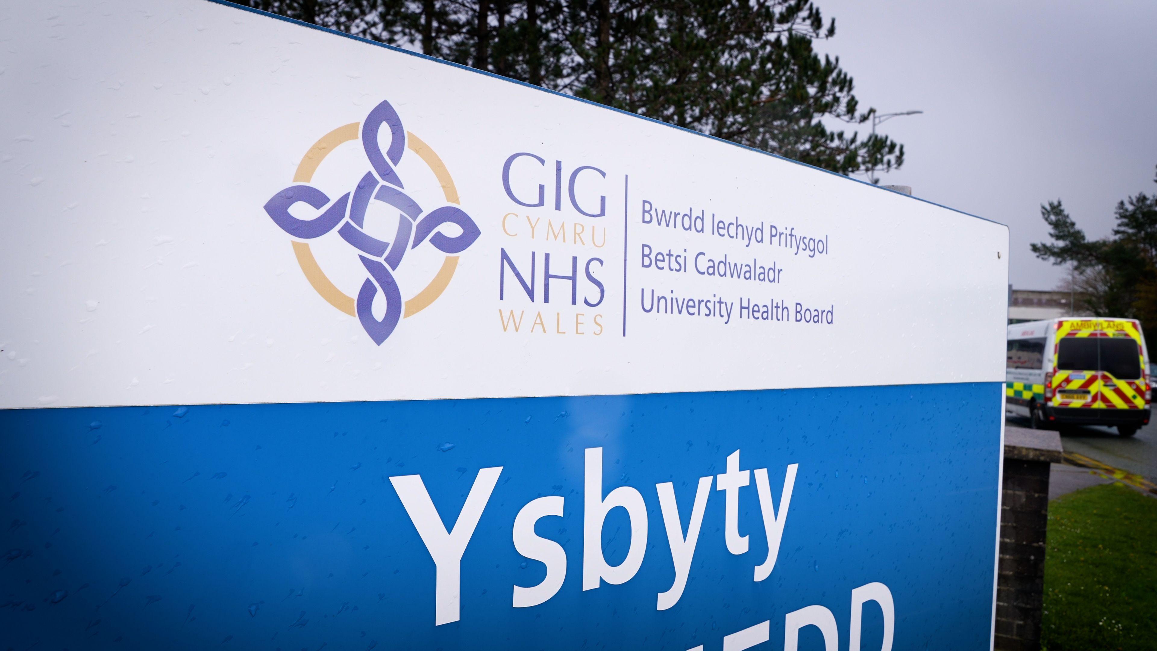 Arwydd Bwrdd Iechyd Prifysgol Betsi Cadwaladr. Mae'r arwydd yn wyn ac yn las ac mae abiwlans yn y cefndir.