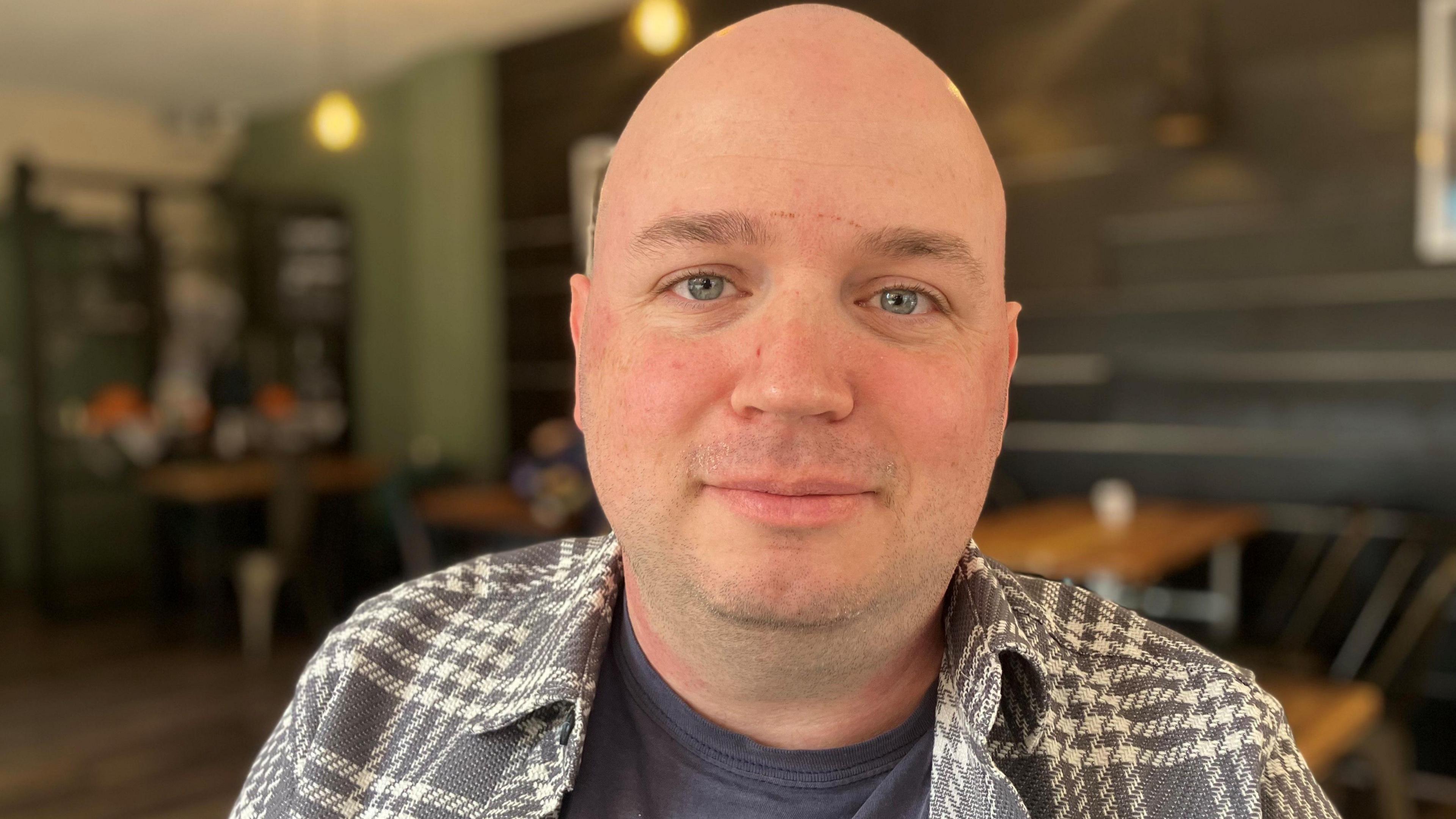 A man with a chequered shirt over a dark blue t-shirt, smiles slightly as he sits in a cafe.