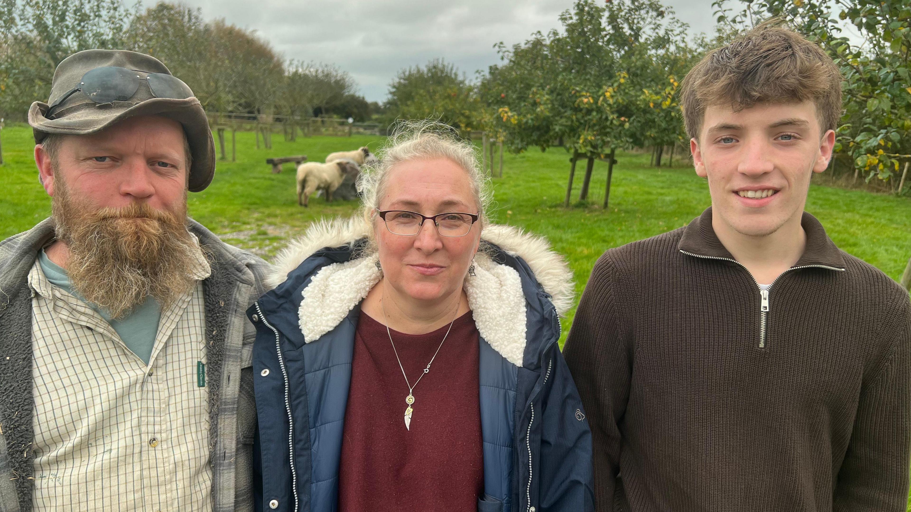 Dave, Kathrin and Seb Kirk smile into the camera, with their field with sheep and trees behind them. 