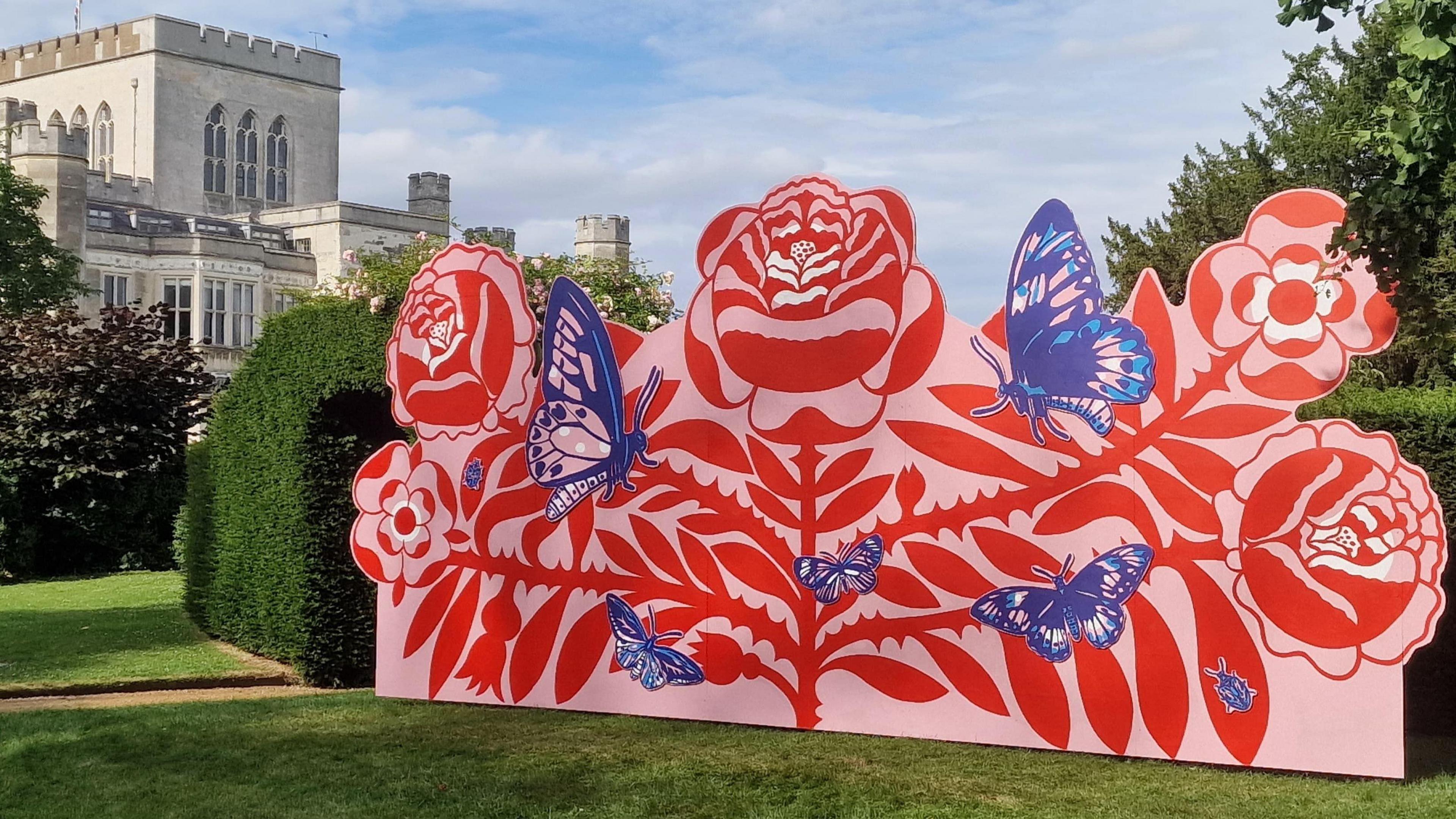 A big piece of art. It is red, pink and blue. Red flowers with blue butterflies have been painted on to a pink canvas.