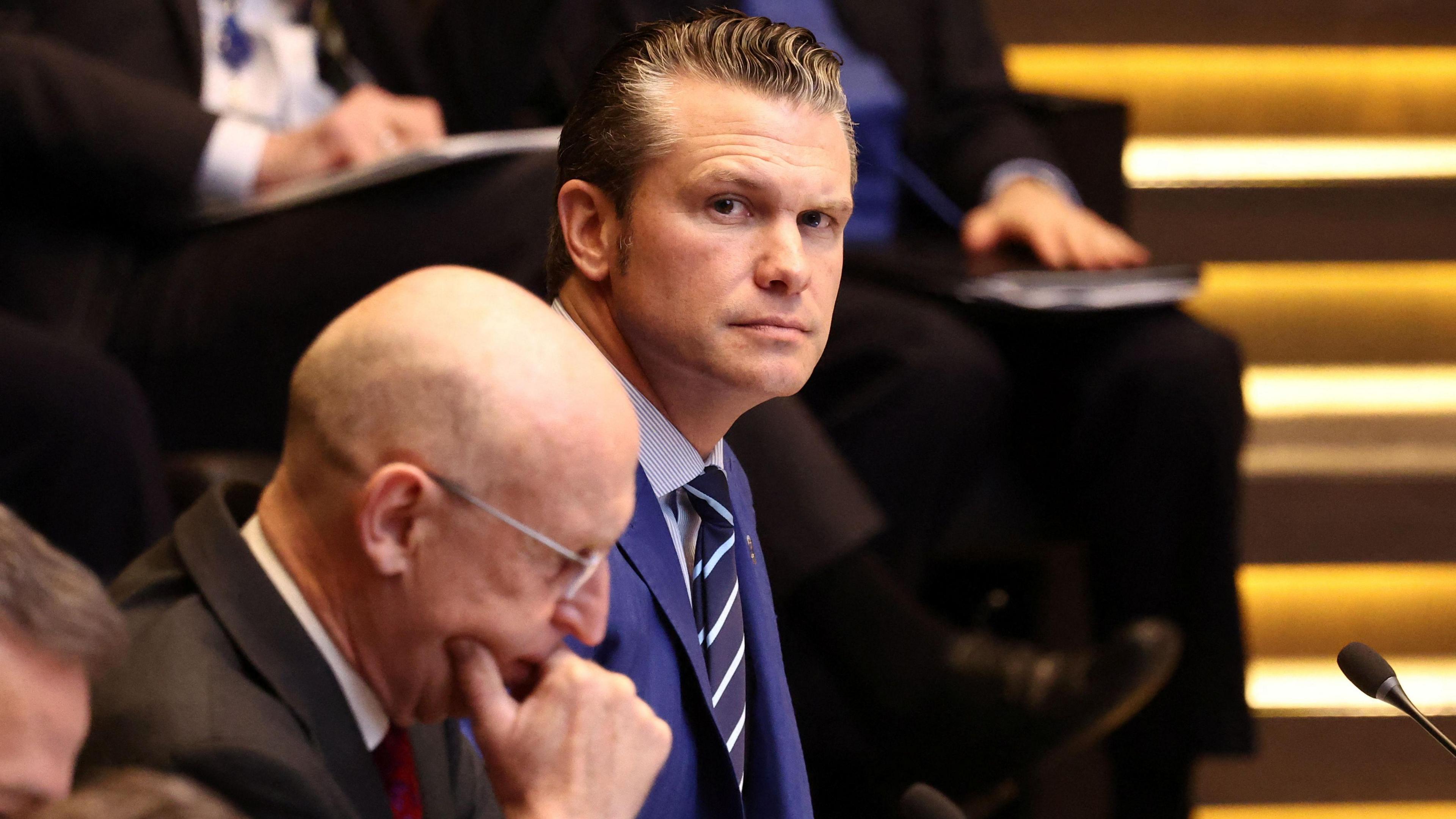 US and UK Defence Secretaries Pete Hegseth and John Healey are pictured next to each other at Nato's HQ in Brussels. Hegseth, with slick-backed greying brown hair looks into the camera, while John Healey, who wears glasses and is bald, studies something in front of him