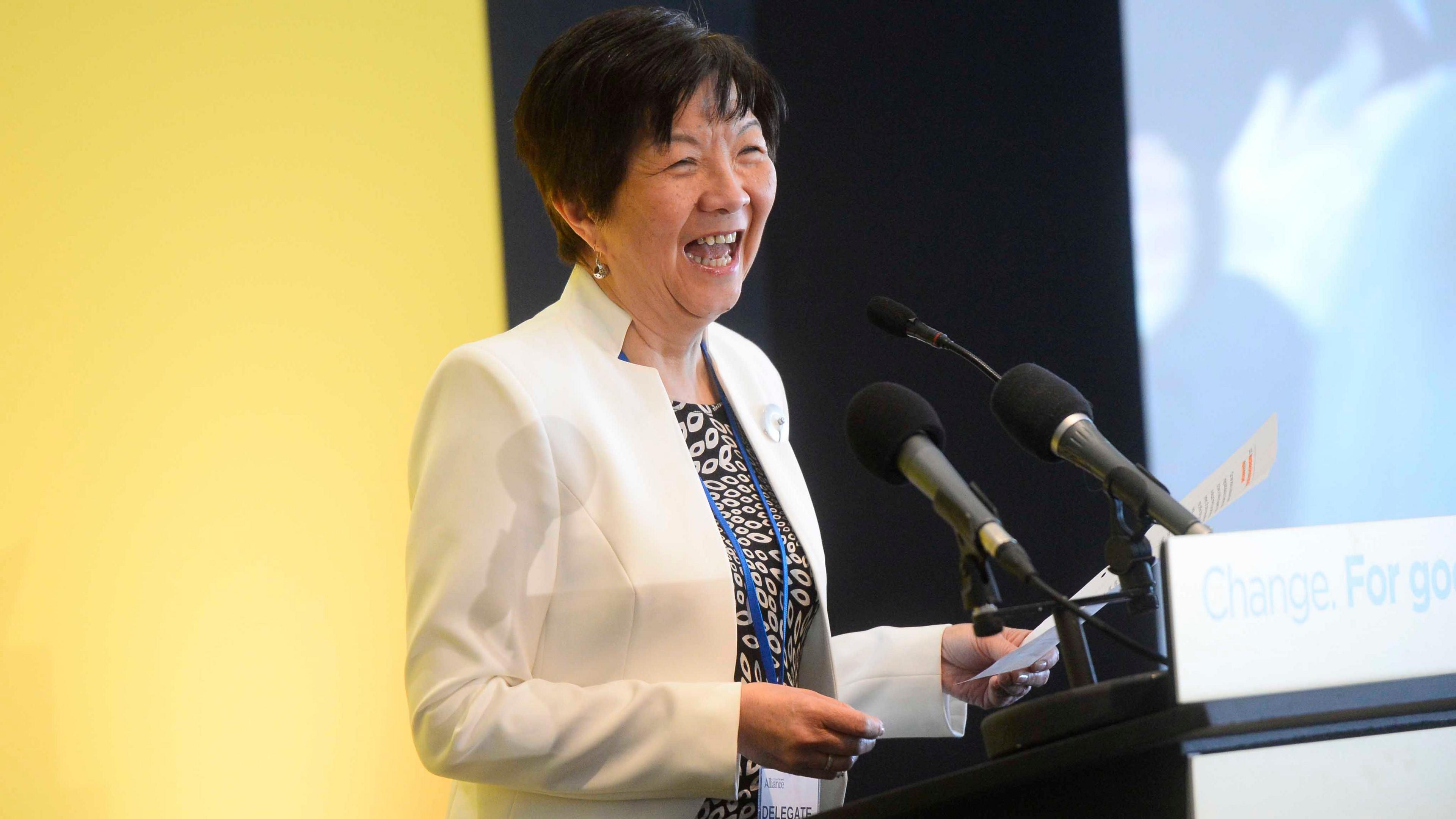 Anna Lo laughing during an Alliance manifesto launch.  She is wearing a white jacket, a monochrome patterned top and is speaking at a lectern. 