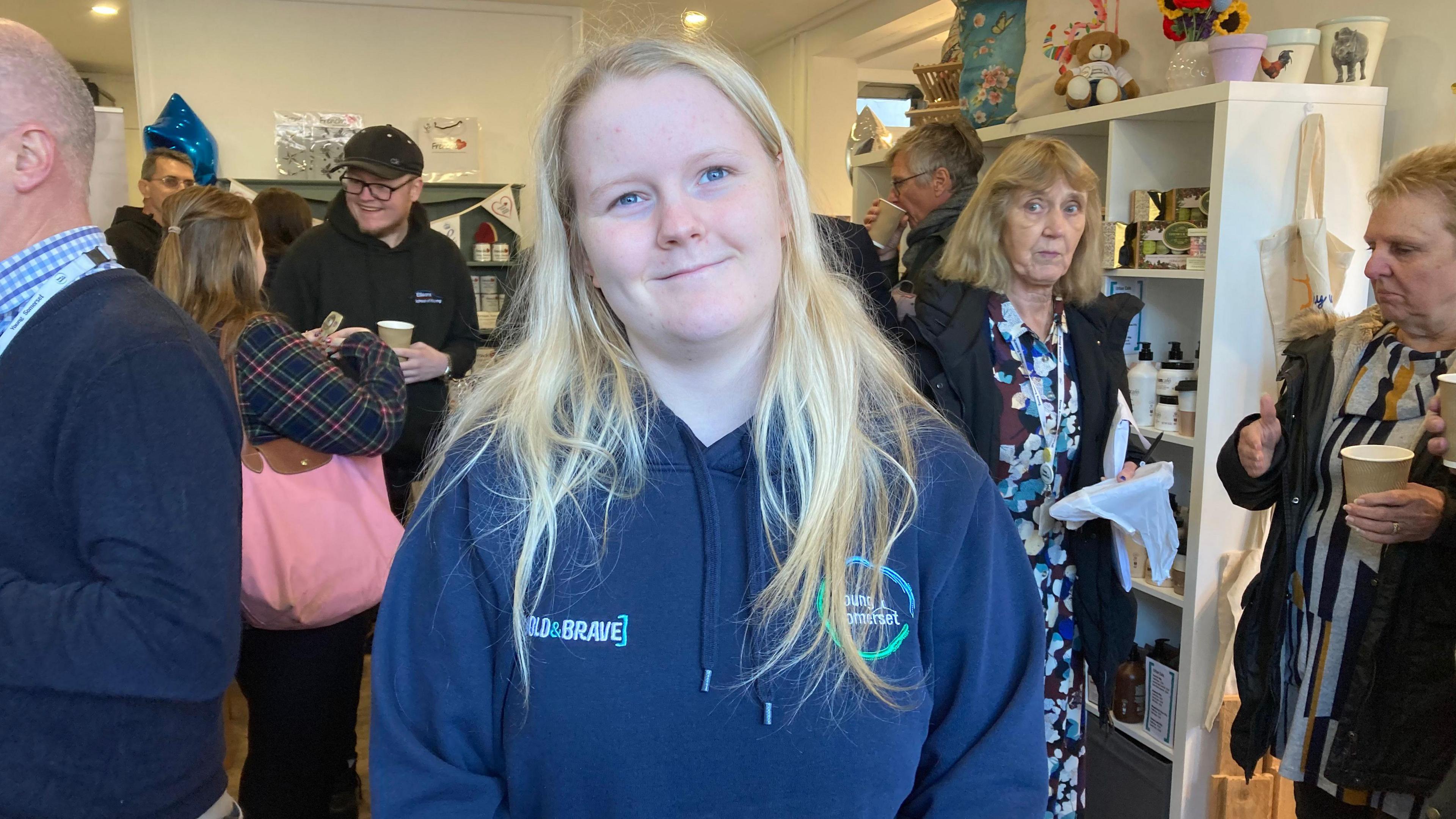 A young woman with long blonde hair looks at the camera with a smile. She's wearing a blue Bold and Brave-branded hoodie. In the background are a number of people chatting inside the shop.