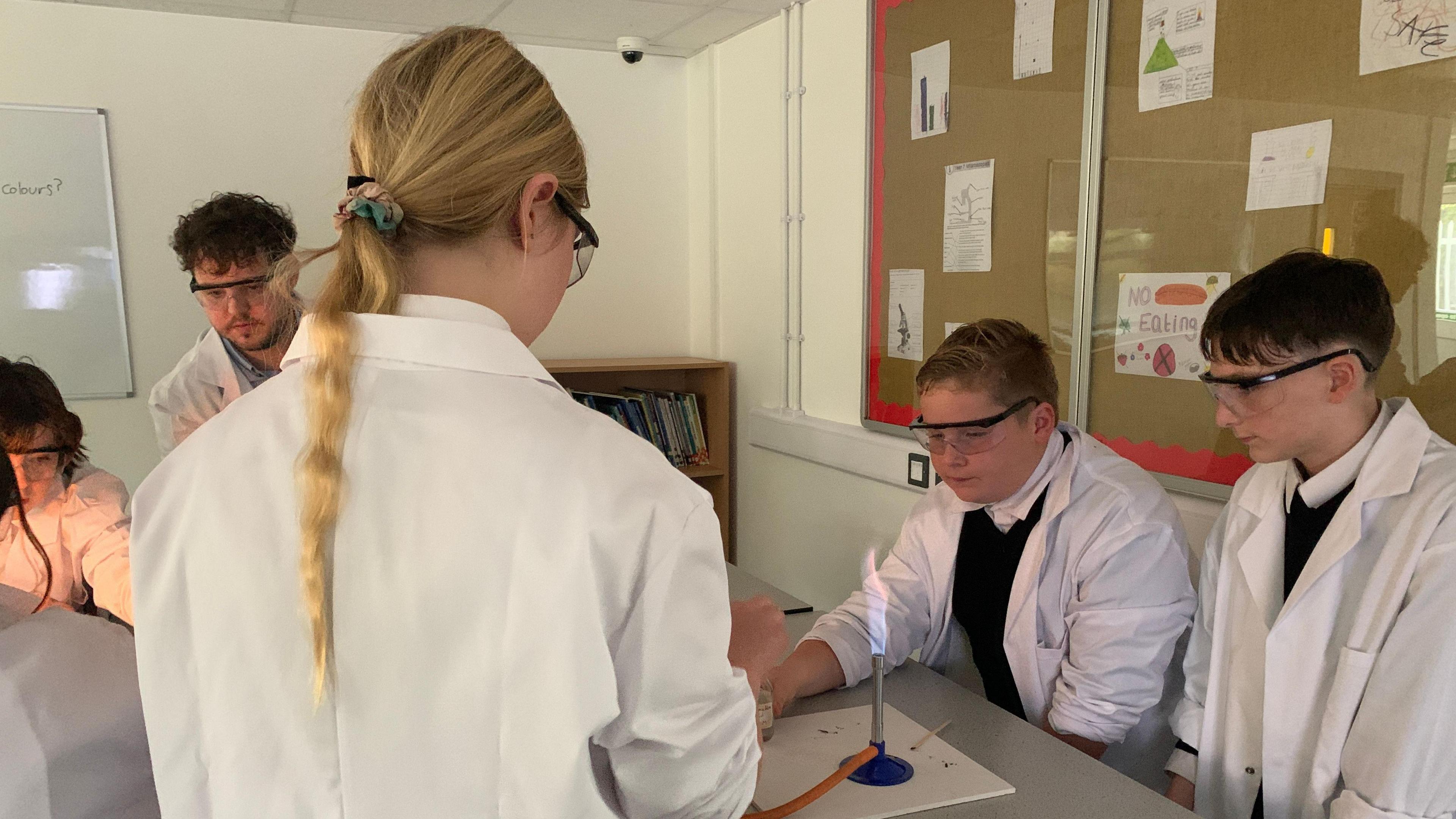 Children gathered round a bunsen burner in white coats watch the changing flames whilst a teacher supervises