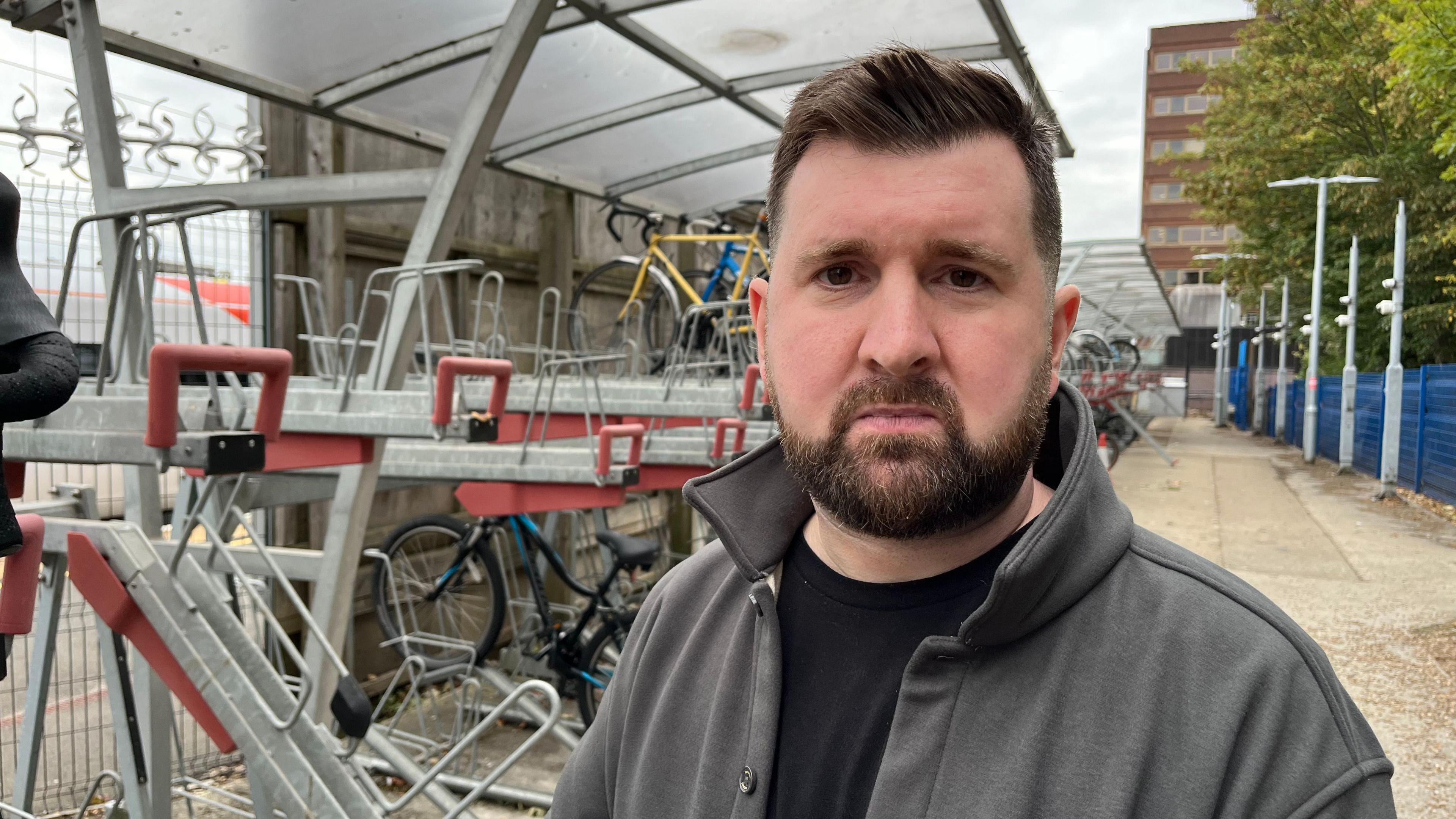 A man in a black top with a beard stands in front of some bike racks