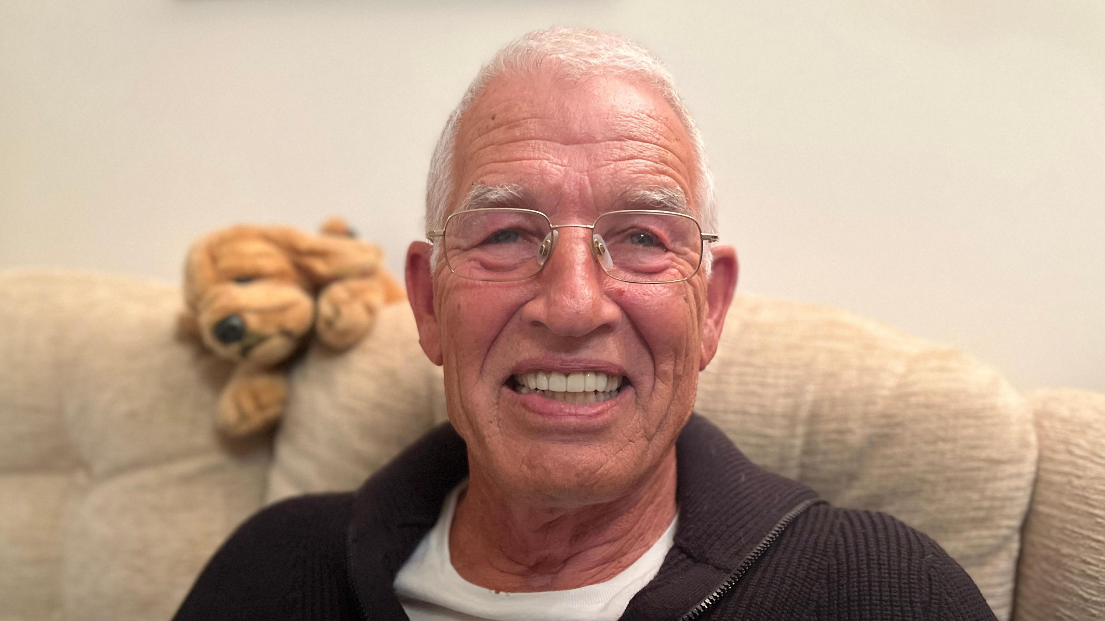 Tim Shepherd grins at the camera while sitting on a sofa. He has short, white hair and wears square, thin-framed glasses, a white T-shirt and a dark, knitted, zipped top.