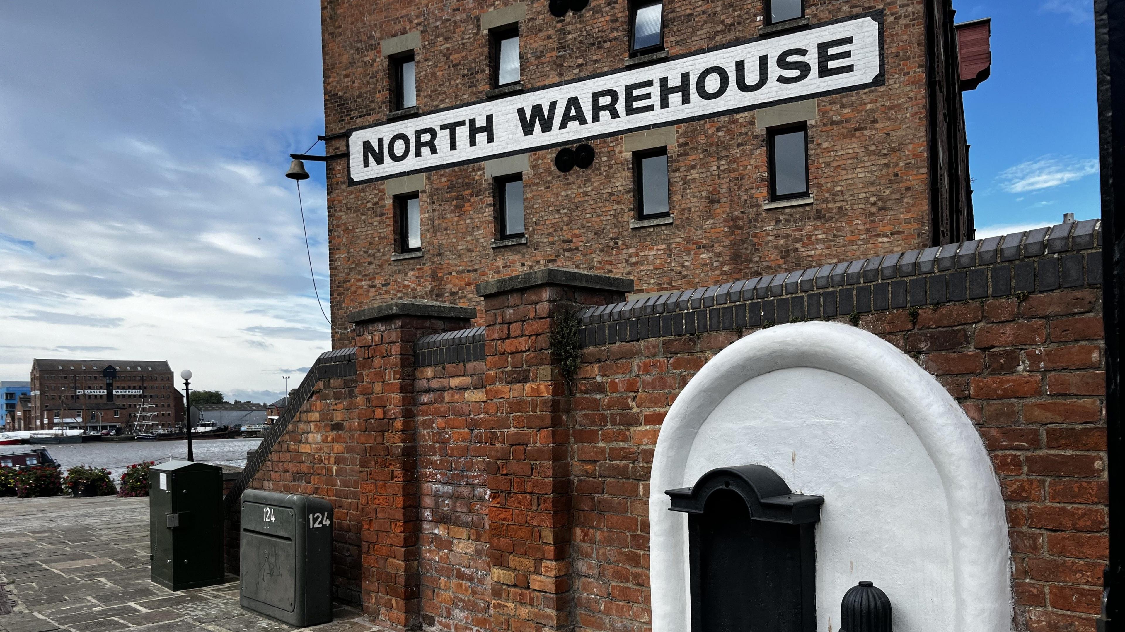 A wide shot of the top of the fountain, looking over the wall towards a large brick building with a painted sign saying 'North Warehouse'. Behind the warehouse you can see the water and boats of Gloucester Docks.