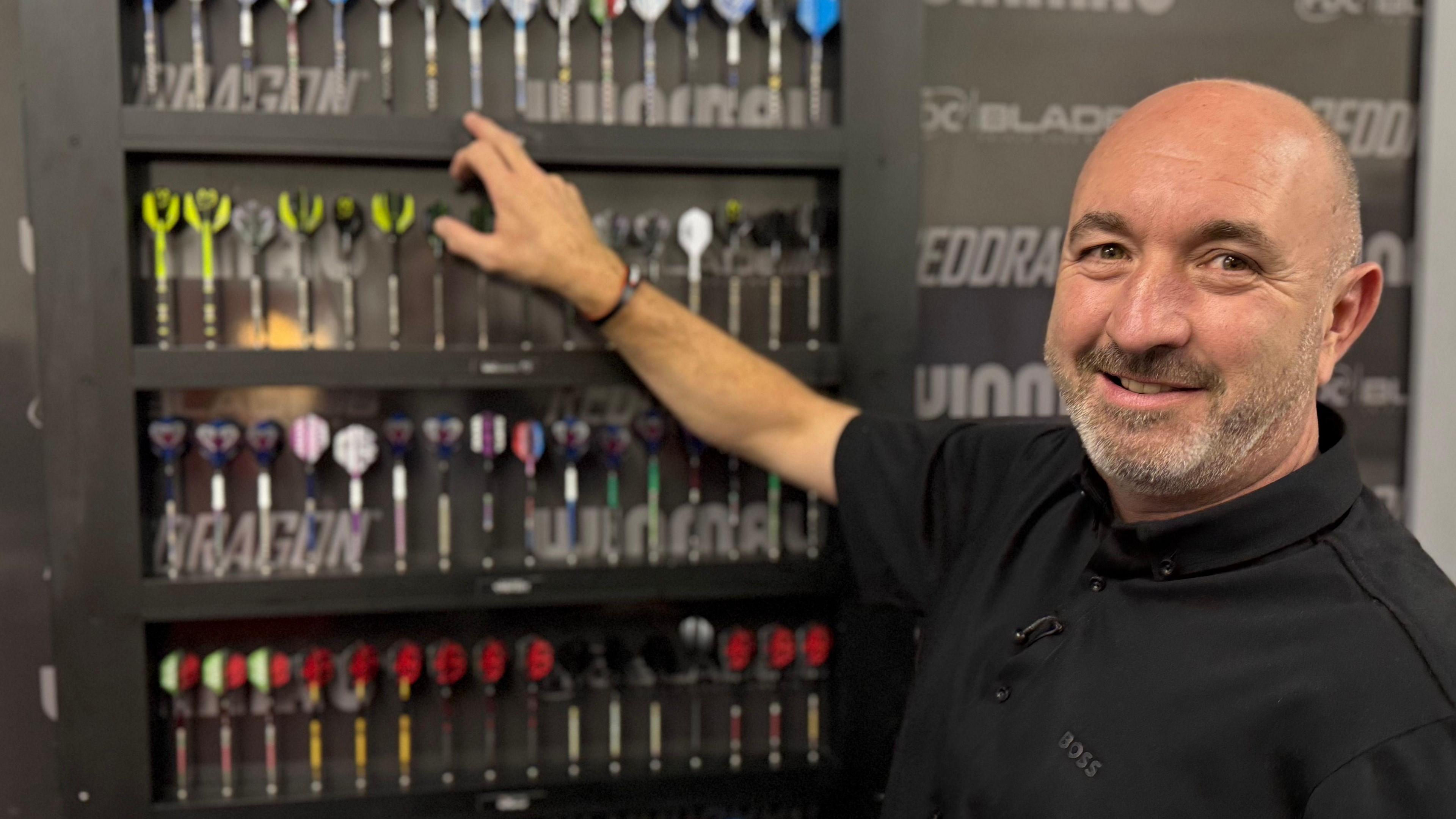 Head of development Lee Huxtable, is pictured next to cabinet which contains five rows of darts. He is pointing at one dart, and is wearing a black polo shirt. Lee is bald with short stubble. 