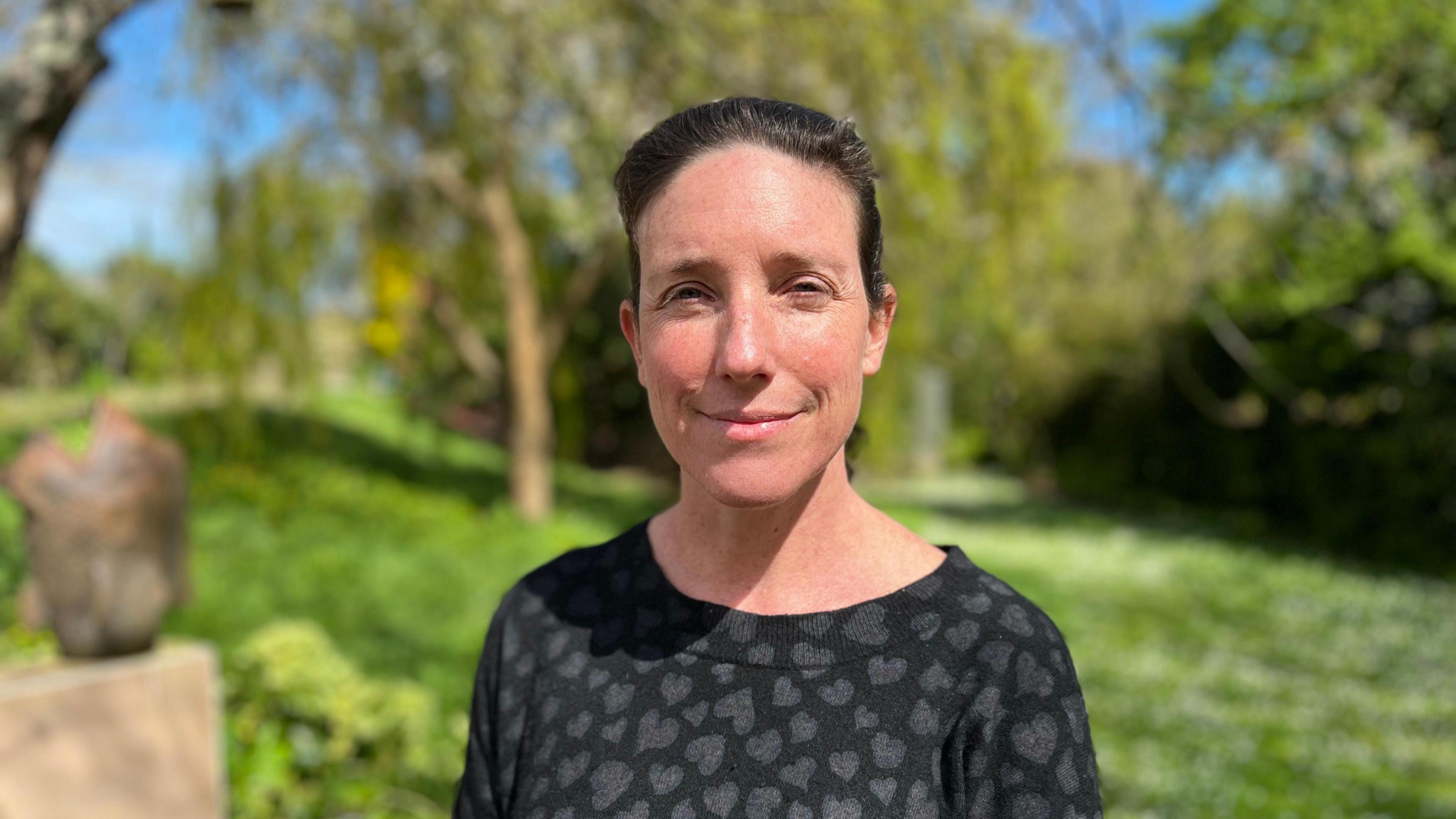 Woman in her 40s in a black jumper top with black hearts on it. She has brunette hair, back in a ponytail. she is stood in a luscious green garden