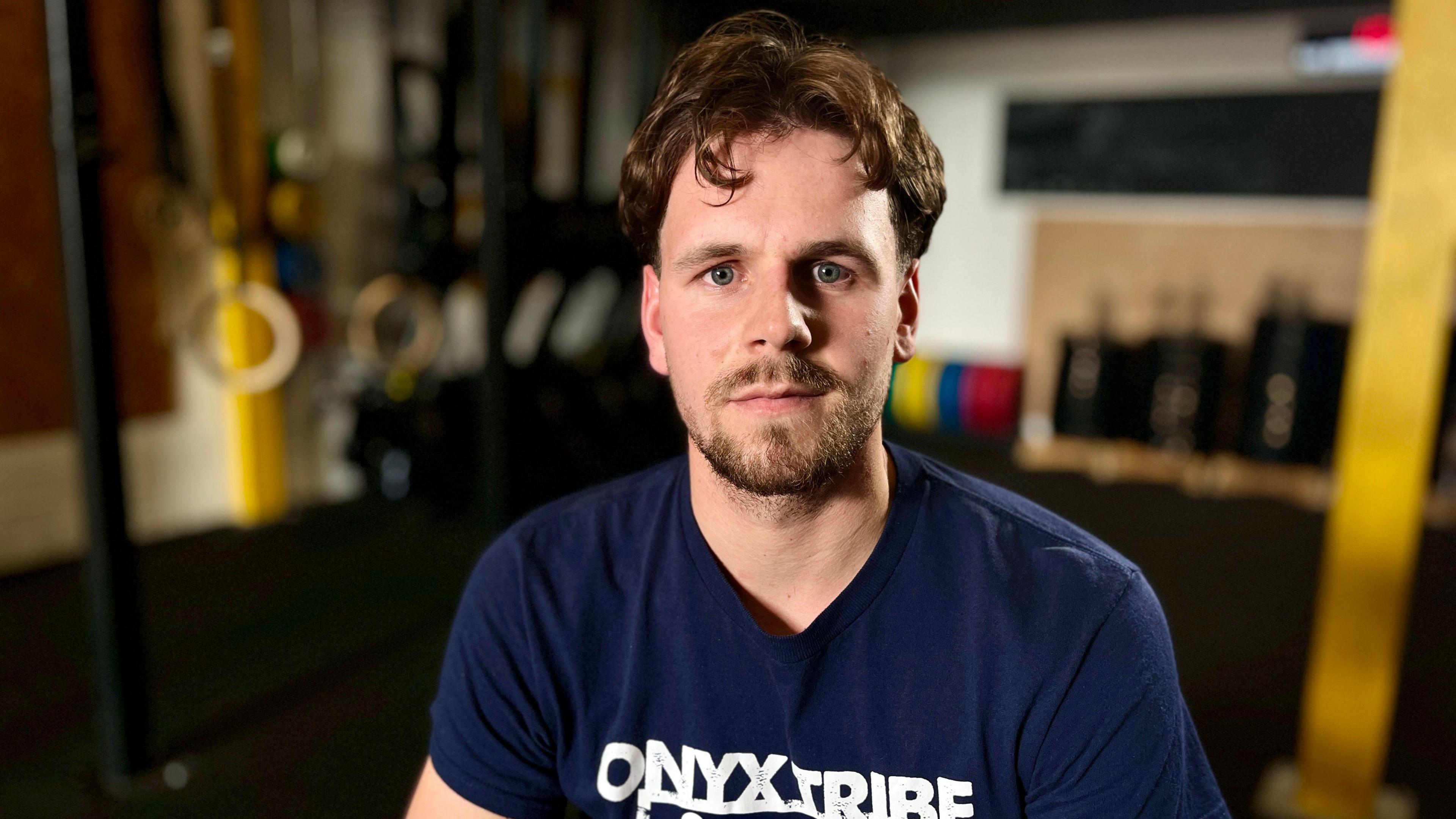 Louis Pearce looks at the camera in the Onyx Tribe Crossfit Gym in Huntingdon. You can see his head and shoulders and he is wearing a navy blue t-shirt. He has brown hair and a short beard. There are gym rings and bar bells in the blurred background.
