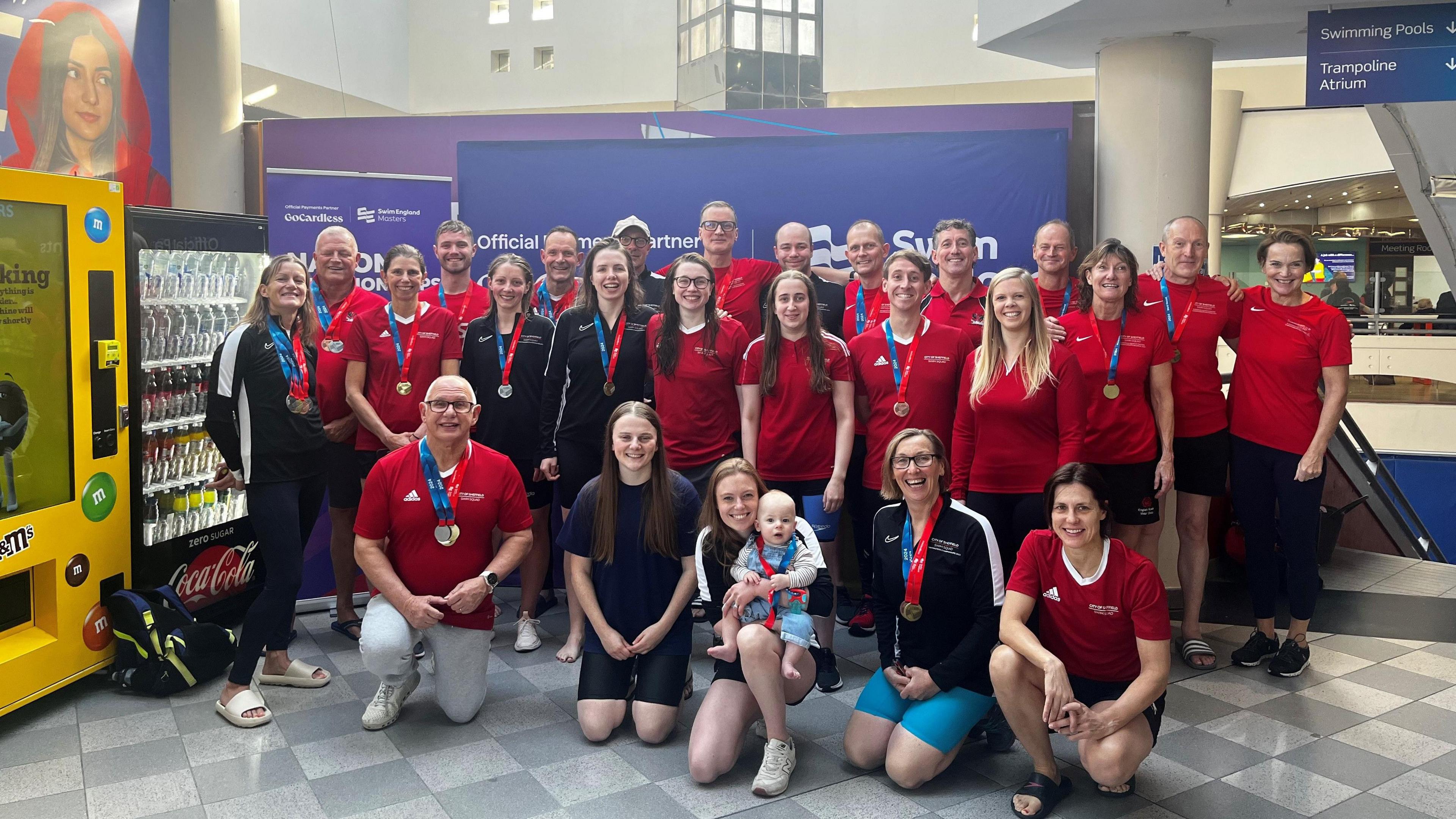 Around 25 people pose wearing red T-shirts and shorts many with medals and in the front a young woman holds a baby wearing medals.