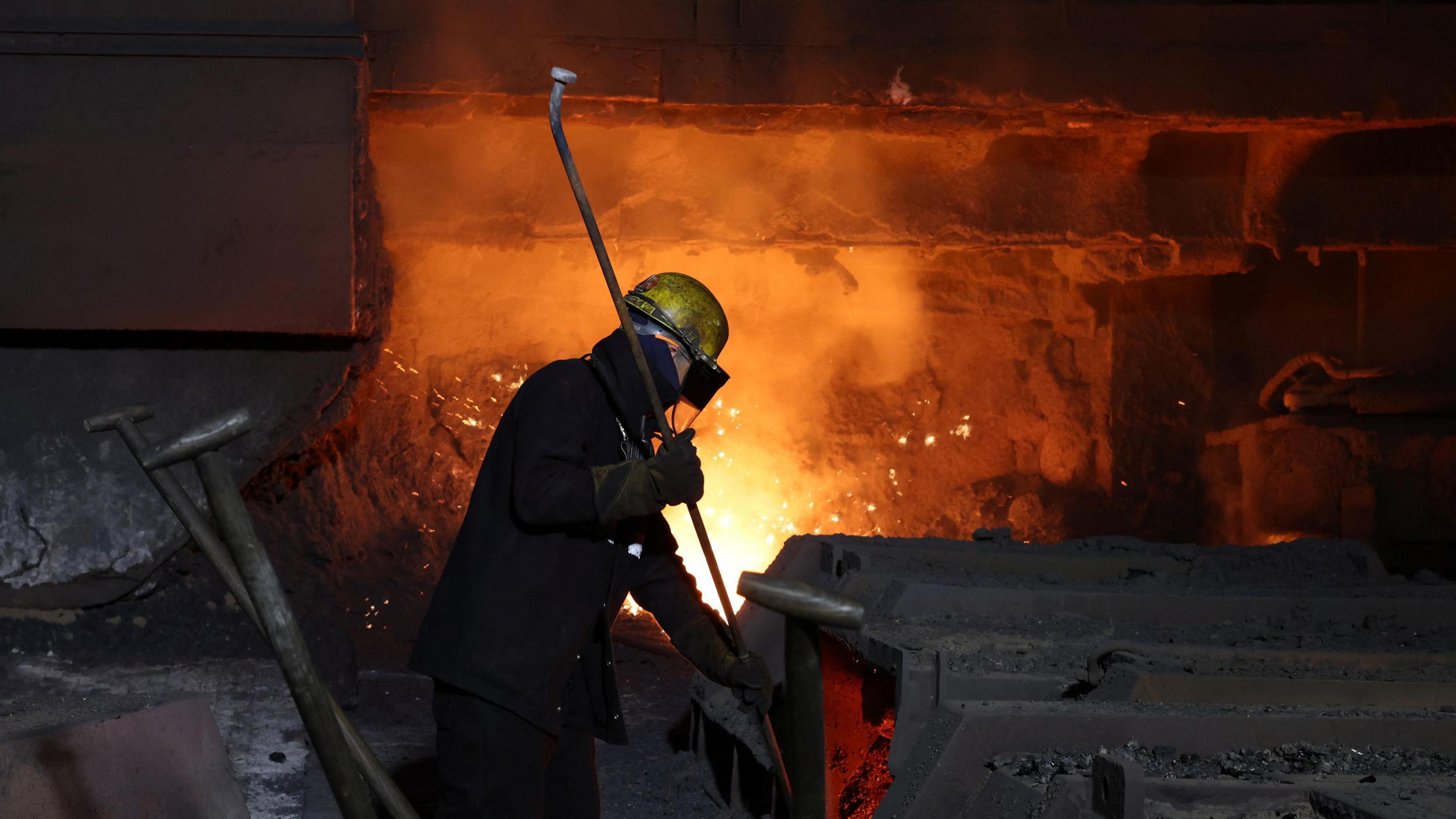 A steel worker at British Steel site in Scunthorpe in April 2025 - the picture is largely dark, which dramatically frames one worker standing in front of a furnace, his outline lit up by the furnace behind him - he is wearing a protective helmet and goggles, and holding what looks to be a long pipe