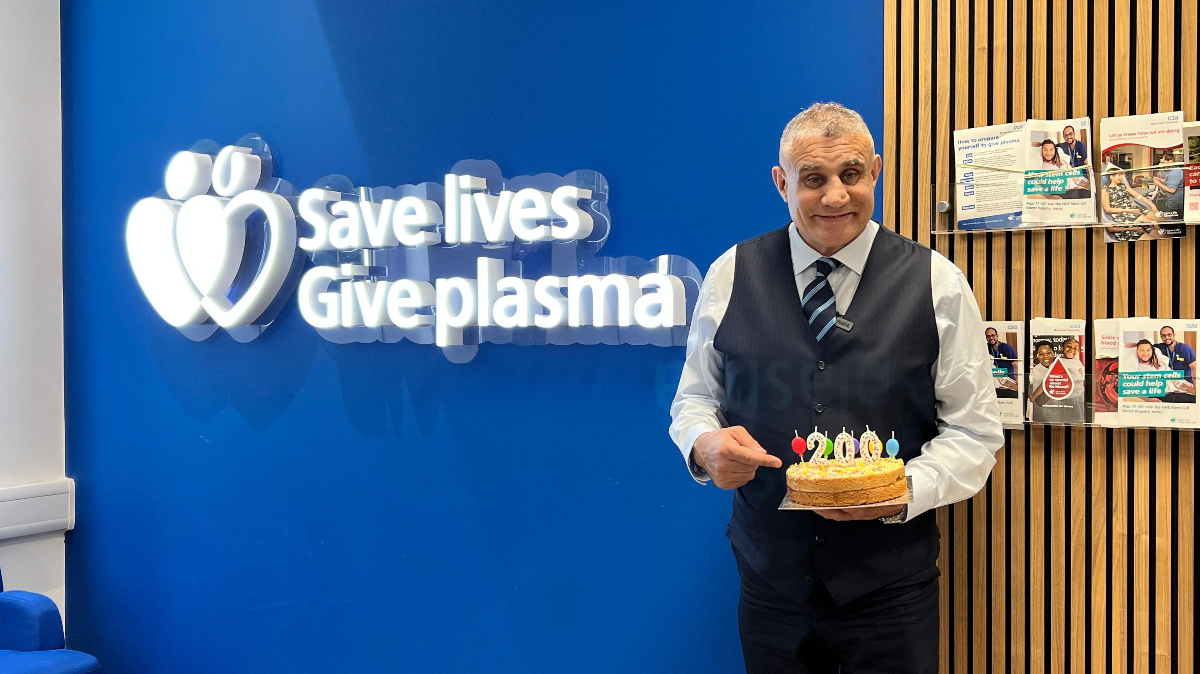 An older man who is wearing a shirt, waistcoat and suit trousers holding a cake. He is posing for a picture in front of a blue wall that had a white sign which reads "save lives give plasma". He is smiling.