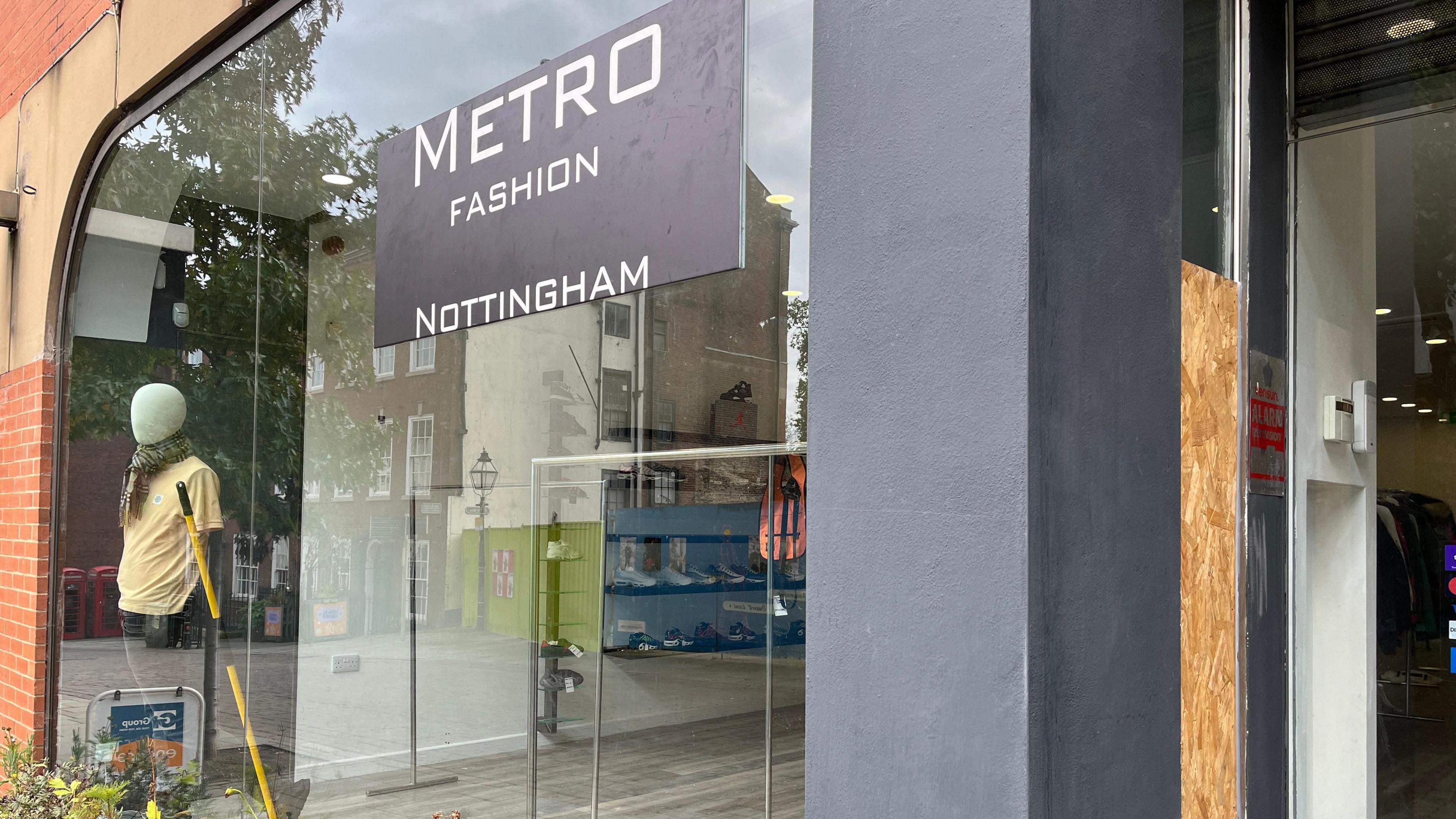 Shop frontage of Metro Fashion showing a large window and doorway, where part of the doorway glass has been boarded over