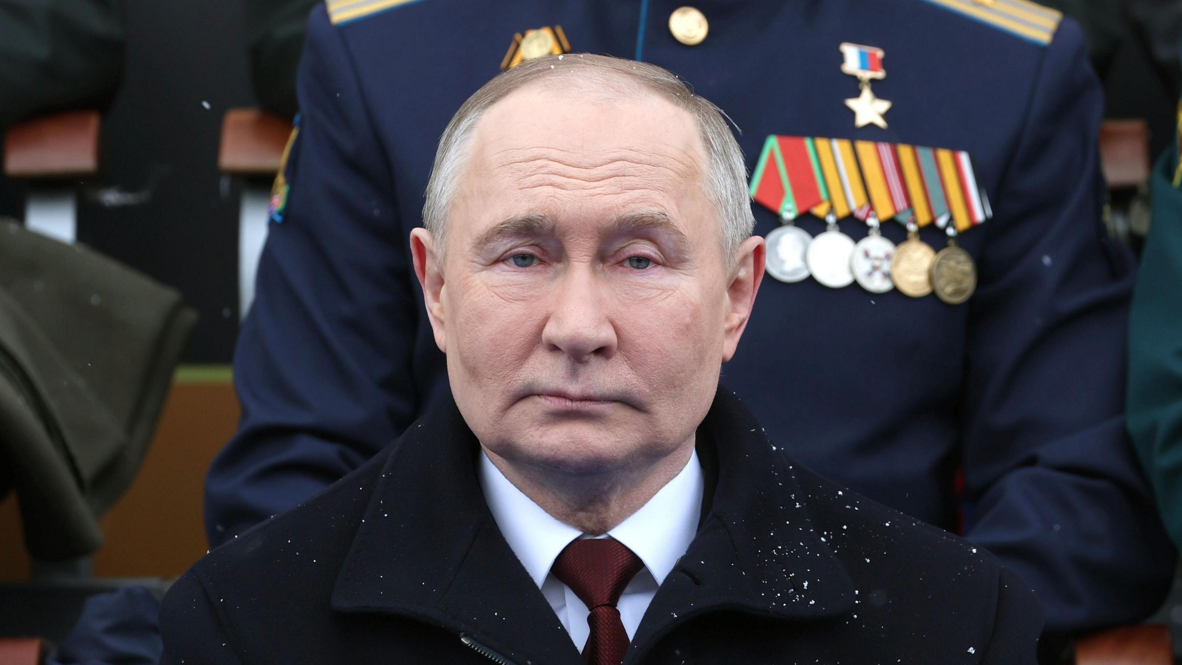 Vladimir Putin stares straight into the camera at a military parade in Moscow, with a dusting of snow on his black overcoat