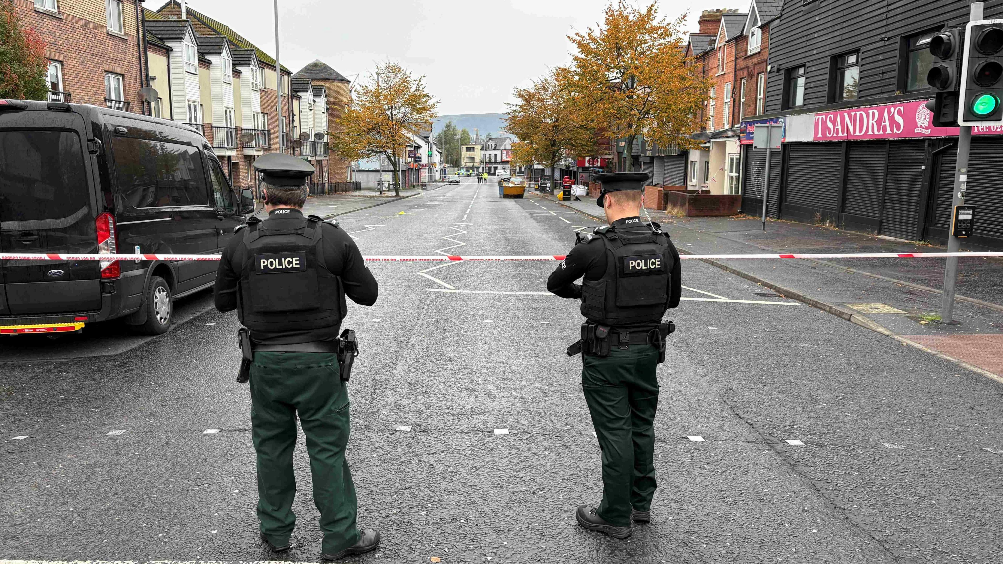 A road which has been cordoned off with red and white police tape. On the right, there is a traffic light. There is a black van parked on the left, and two police officers standing in the foreground. 