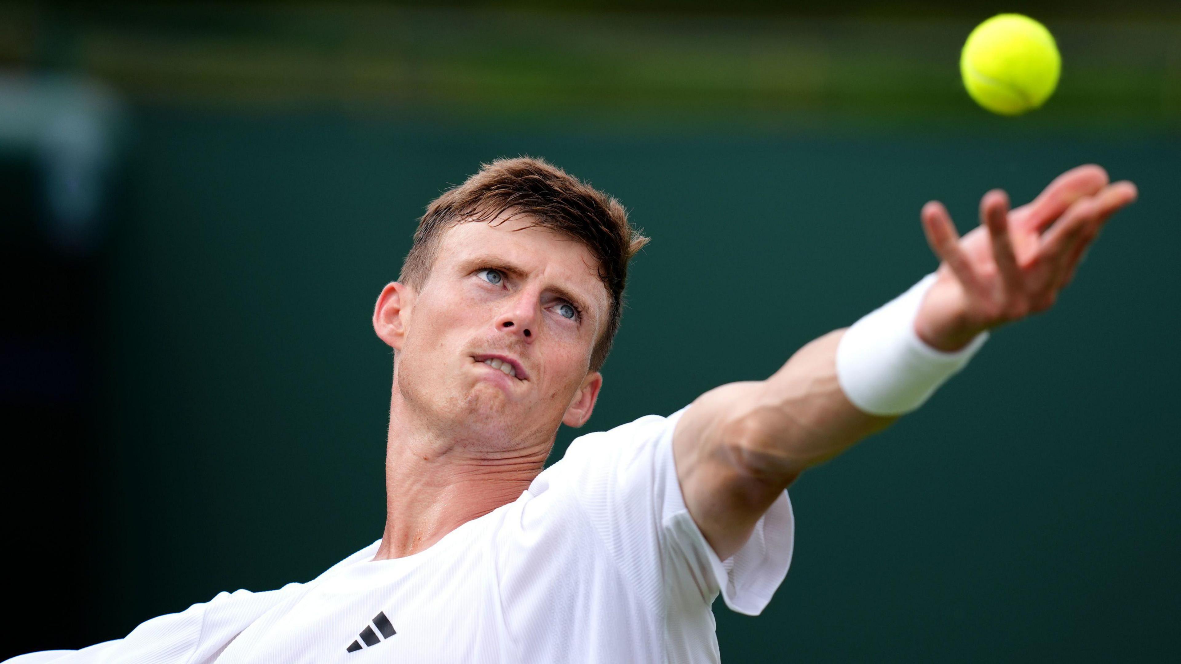 Billy Harris, who has short brown hair and is wearing a white T-shirt and wristband, bites his bottom lip as he throws a tennis ball in the air while serving.