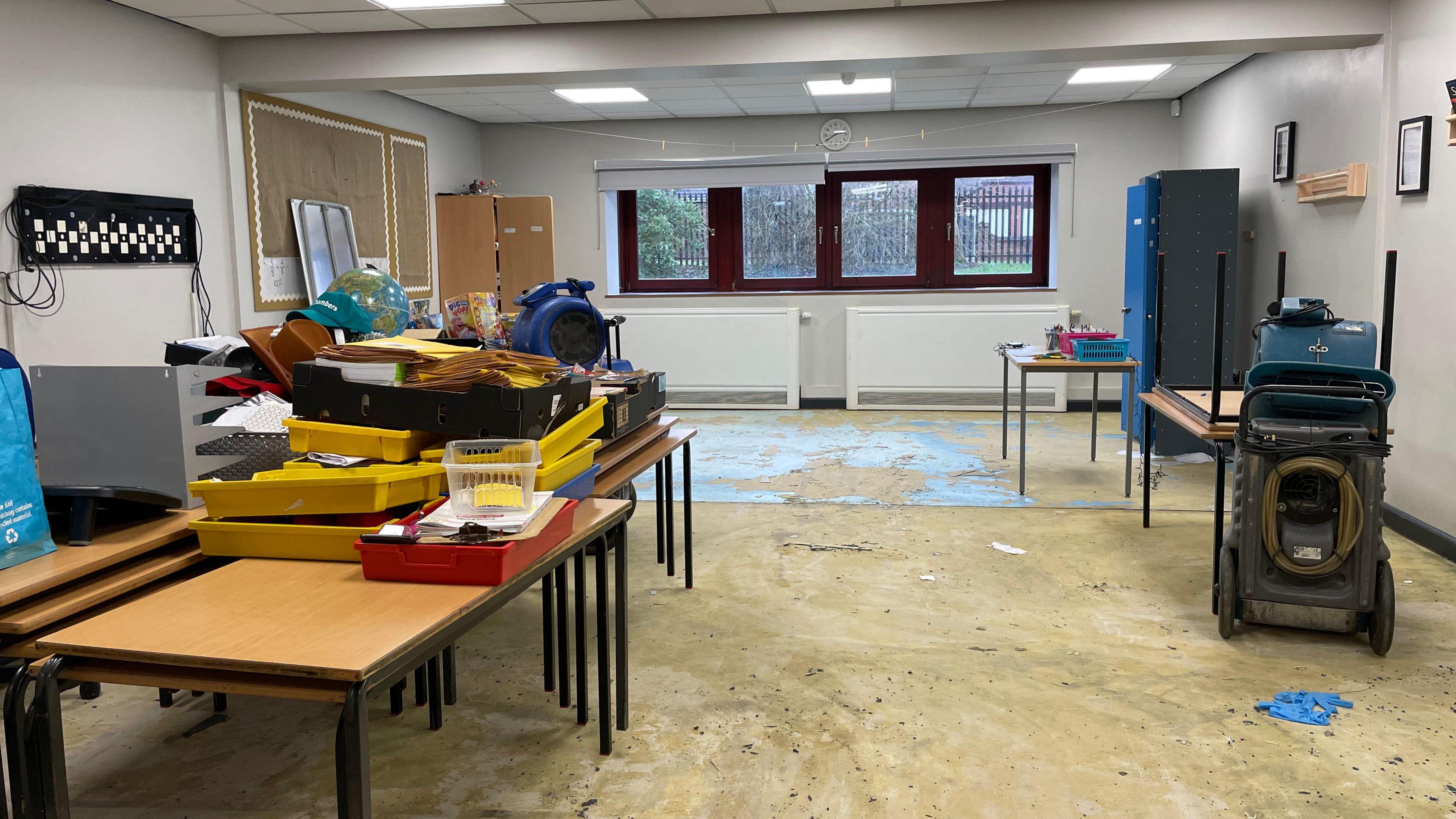 Flooring is ripped up in a school room. Trays and various equipment is stacked on top of tables.