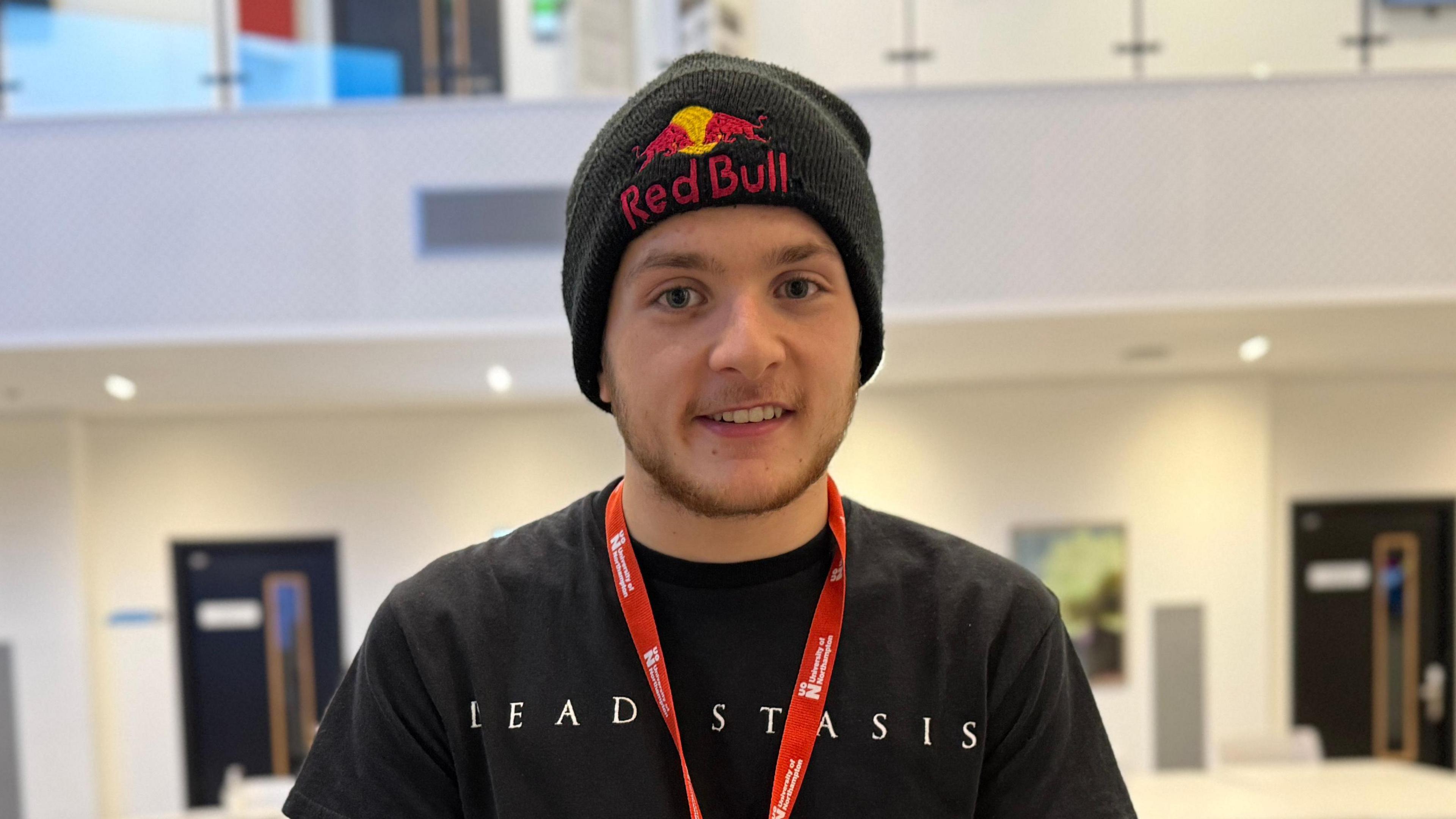 A man looking directly at the camera and smiling wearing a black t-shirt, a Red Bull beanie and a red University of Northampton lanyard. 