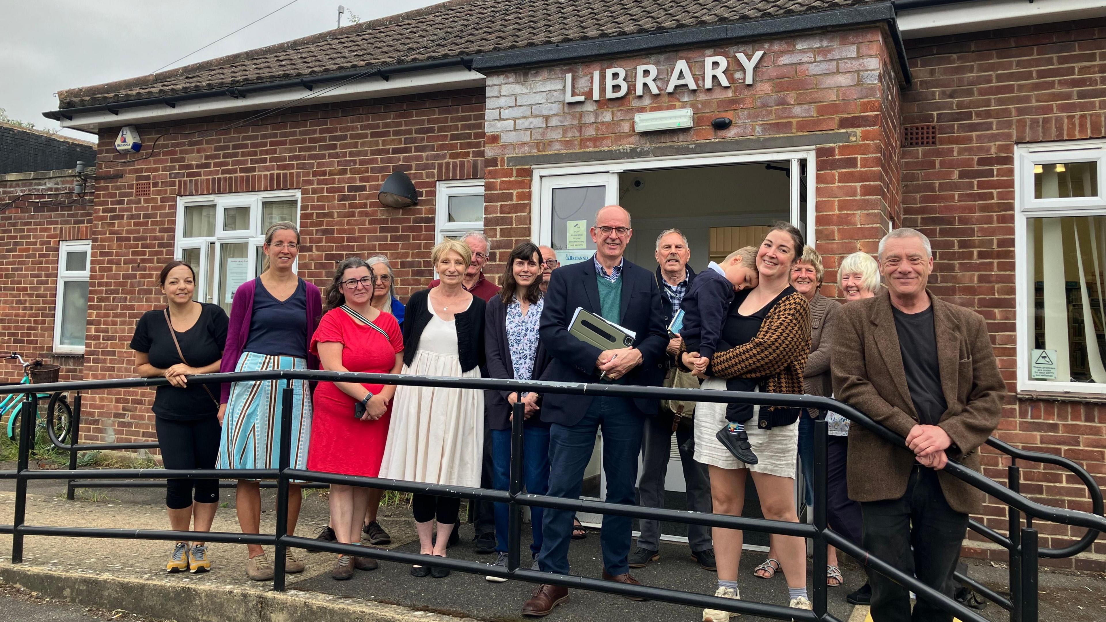 A group of people stand in front of a red-bricked building with "Library" written above the door in large, silver letters