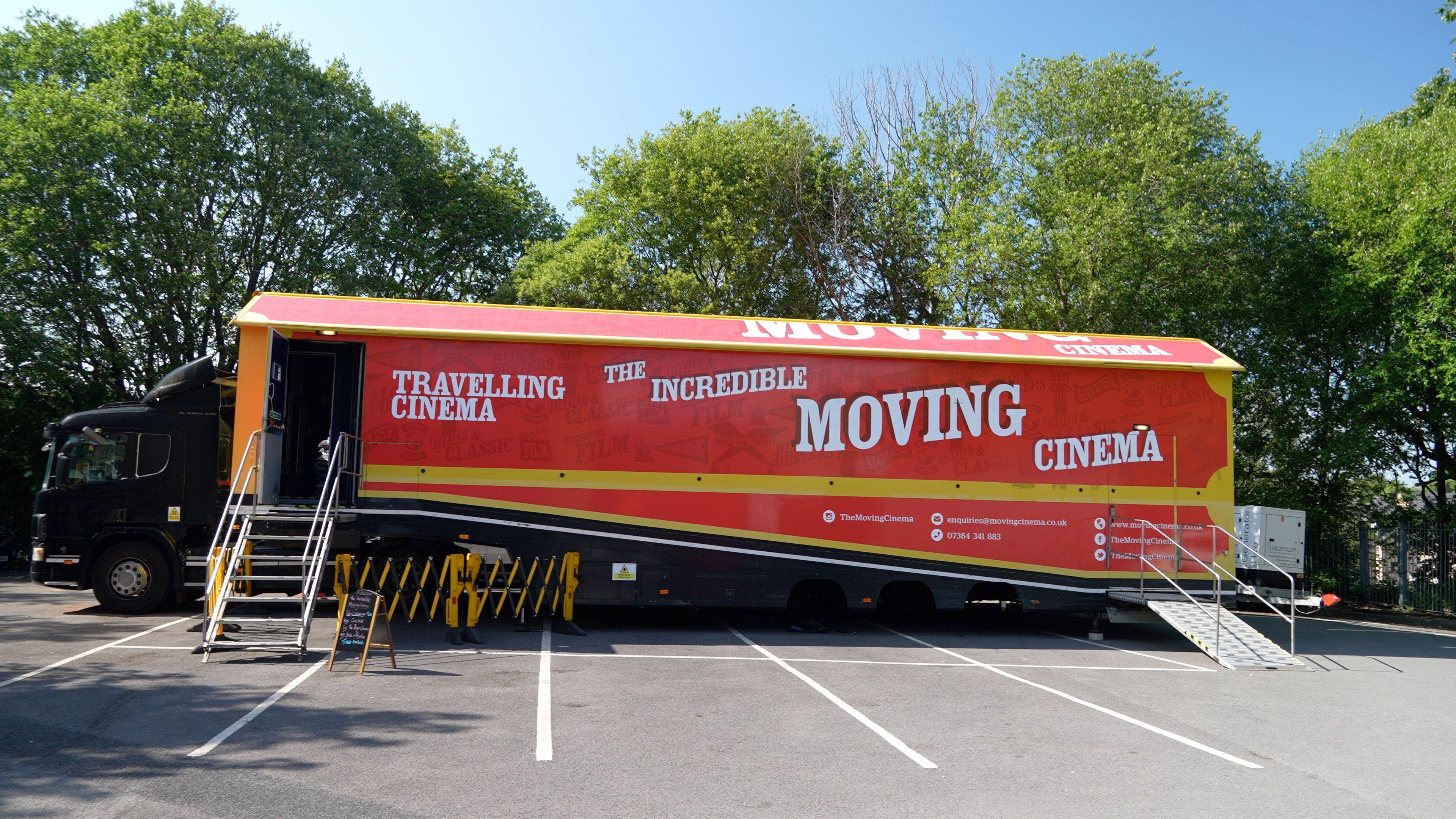 A lorry parked in a car park in front of some trees. The red lorry has the words 'travelling cinema' and 'the incredible moving cinema' on the side. A set of stairs up to a door into the lorry.