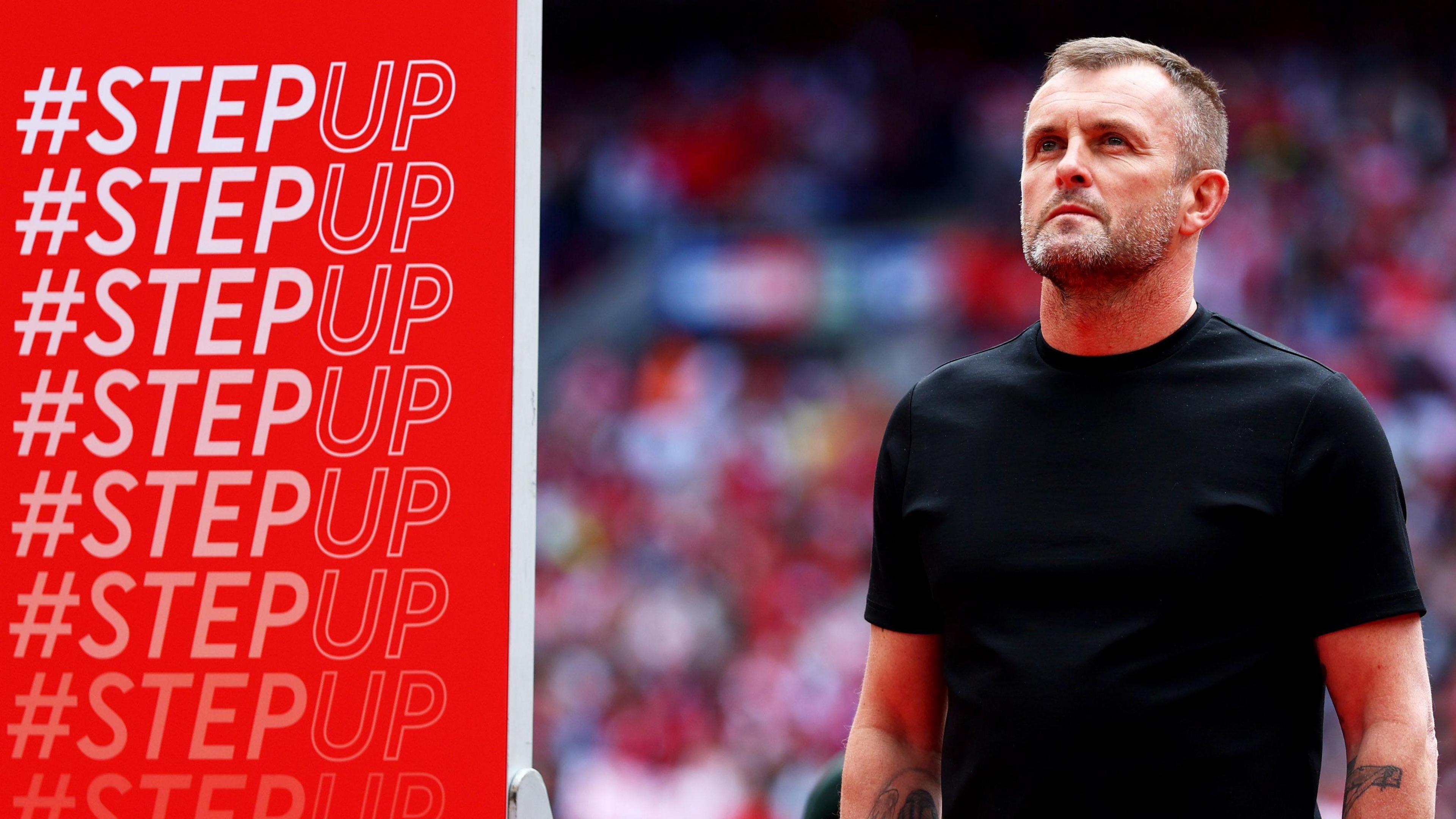 Charlton Athletic manager Nathan Jones stands next to a Step Up banner on the pitch following promotion at Wembley in May